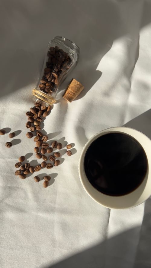 Free Coffee beans spill from jar beside cup of coffee on white cloth in sunlight. Stock Photo