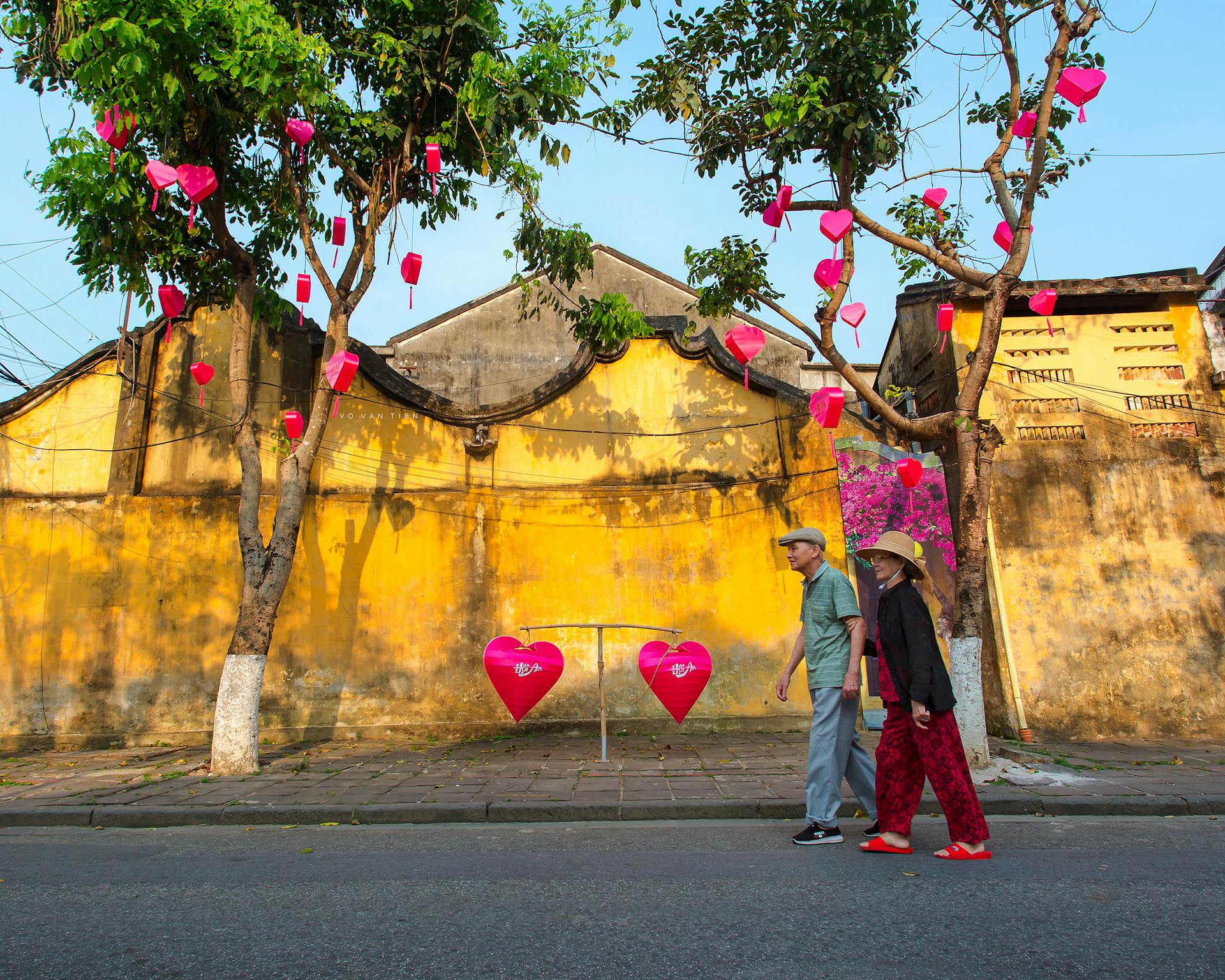 An older couple walks past heart-shaped lanterns on a vibrant street in Hội An, Vietnam.