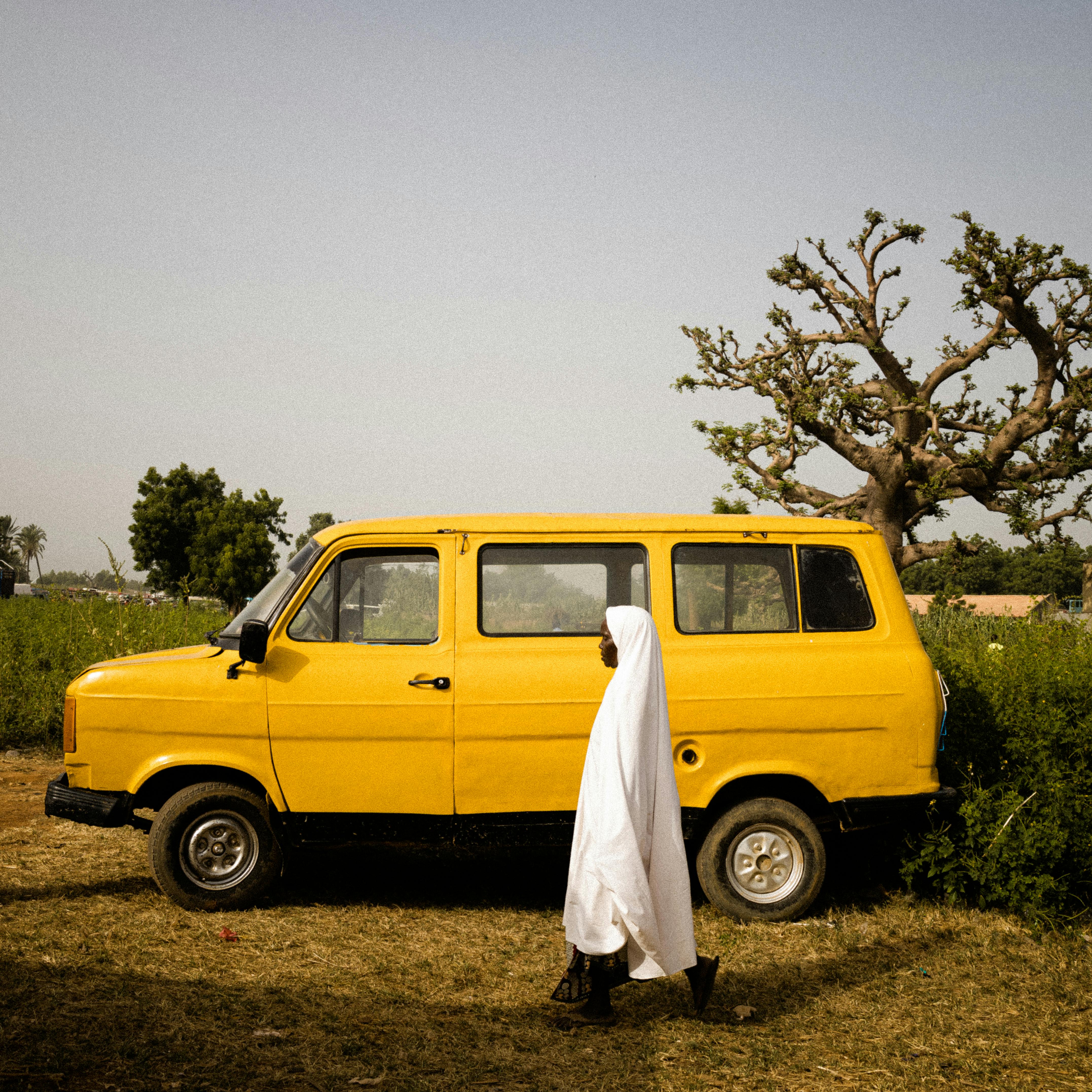 A person in a white robe walks by a vibrant yellow van in a rural landscape.