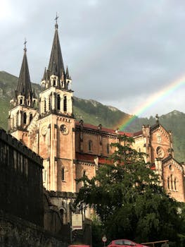 Beautiful view of the Basilica of Covadonga framed by a vibrant rainbow and lush green mountains.