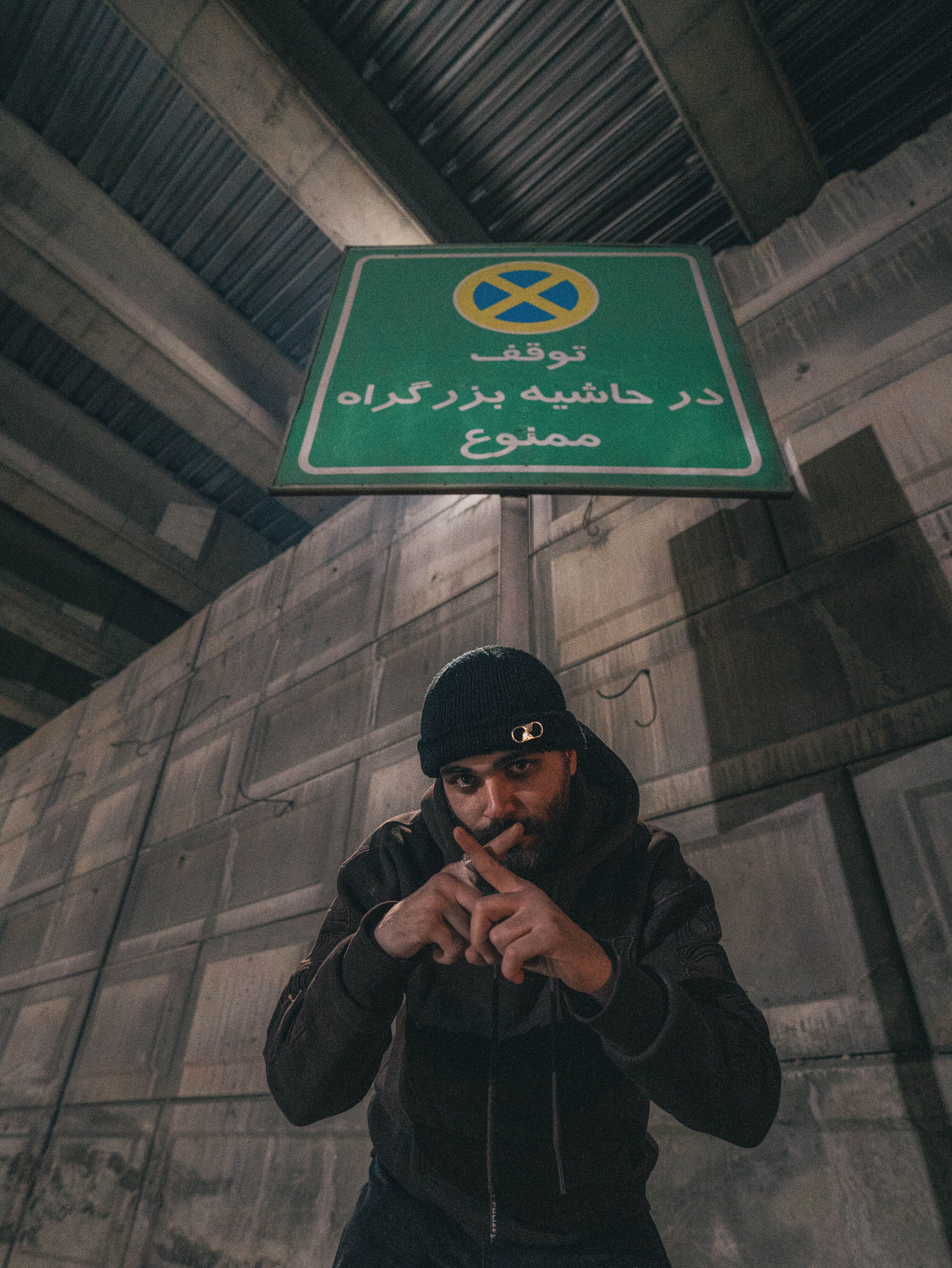 A man poses under a Persian language no parking sign in an urban setting.