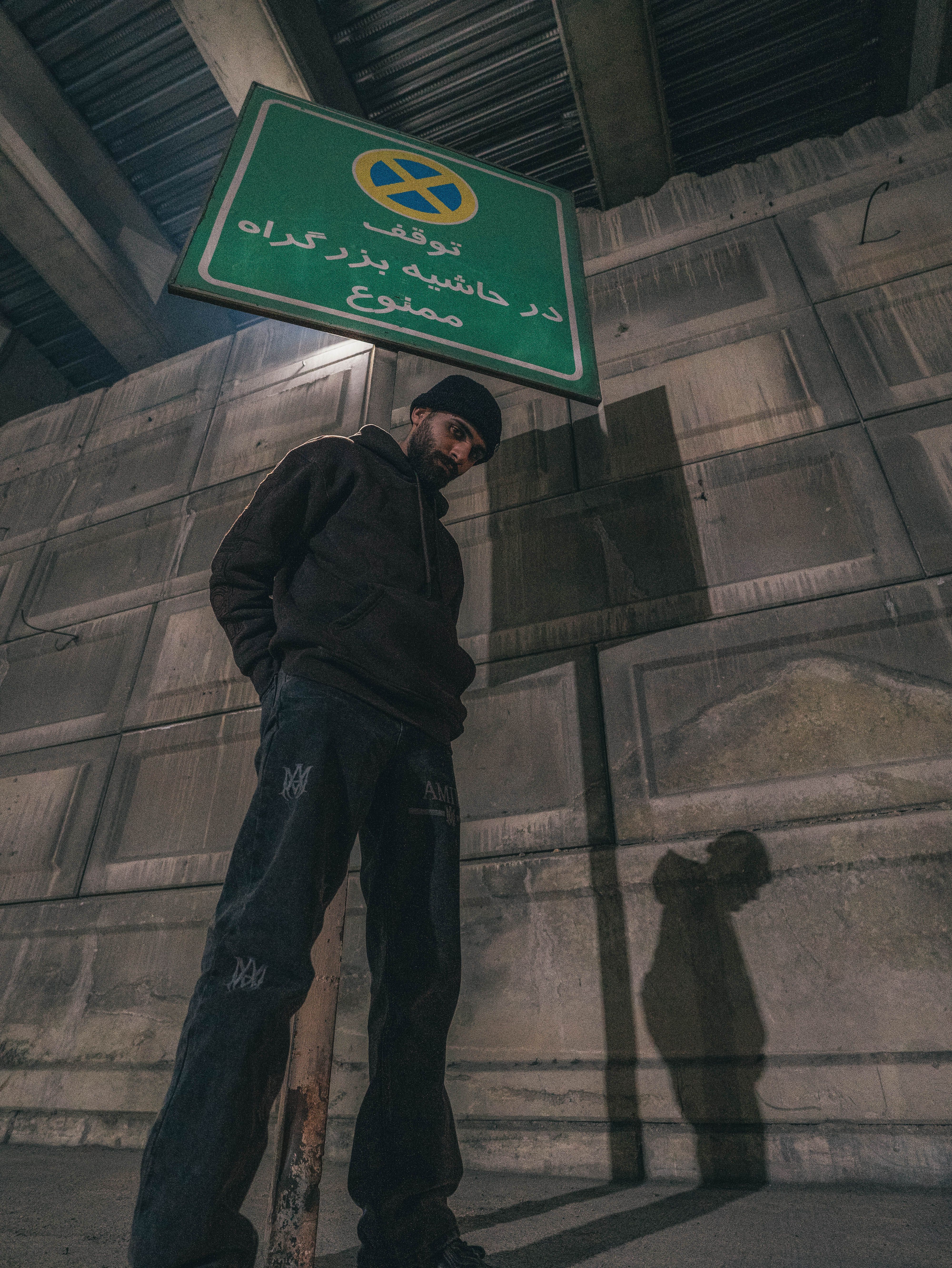 A person stands under a street sign in an urban setting, casting a shadow in low light.