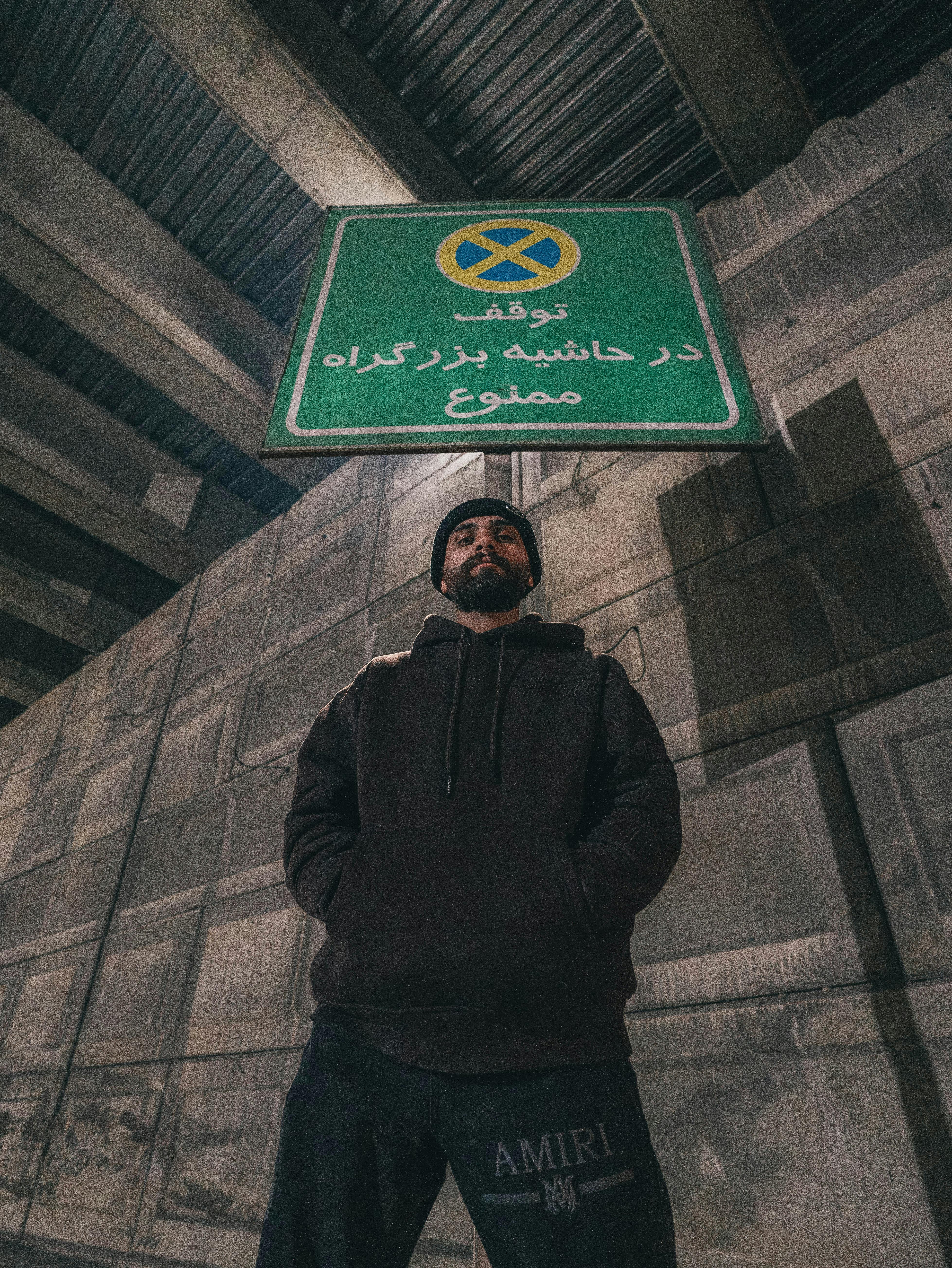 Man standing beneath a green traffic sign in an urban setting, wearing a hoodie.