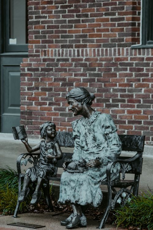 Free Two bronze statues of a child and elder woman sitting on a bench against a brick wall. Stock Photo