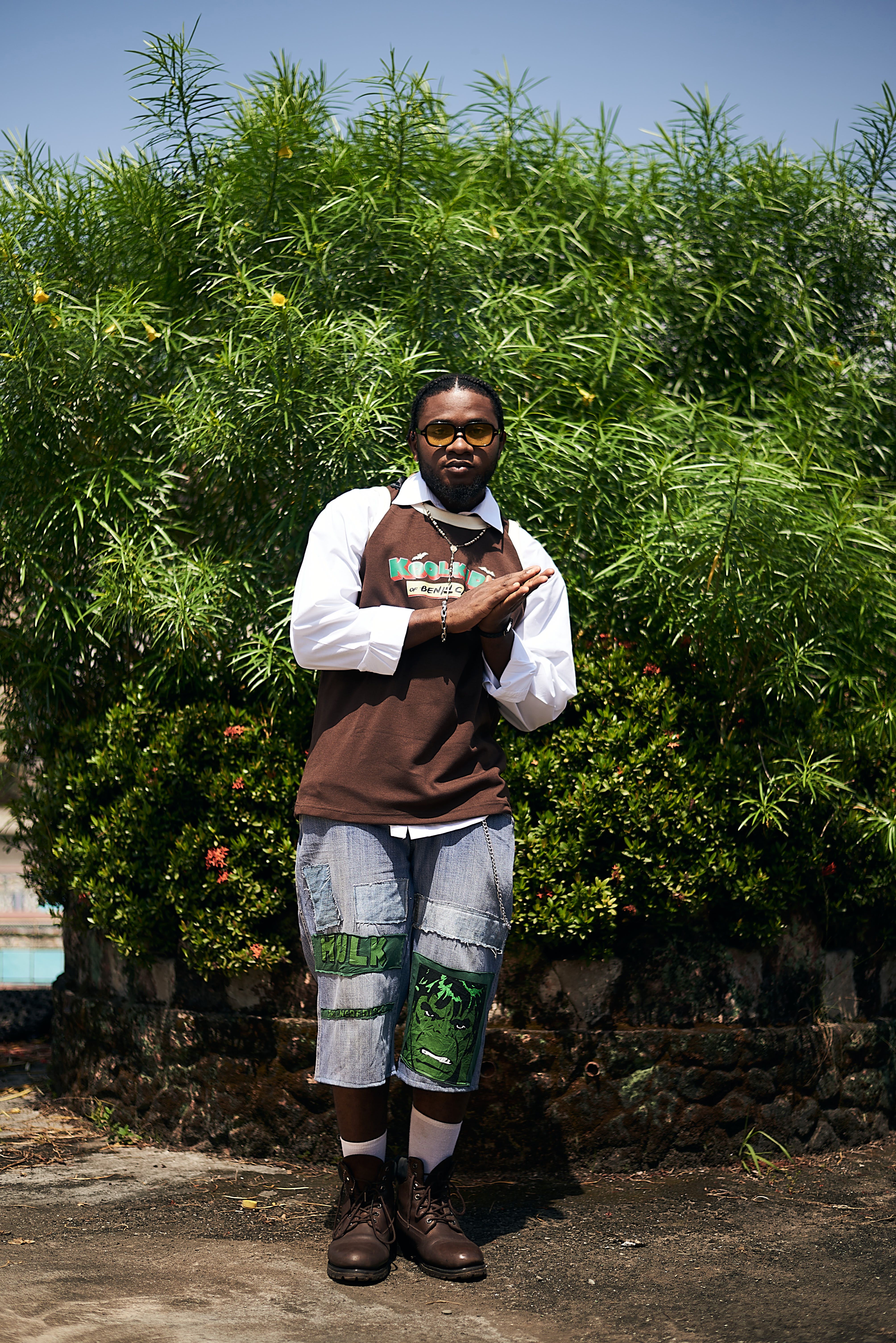 A fashionable adult man poses confidently in front of vibrant green plants, displaying unique style.