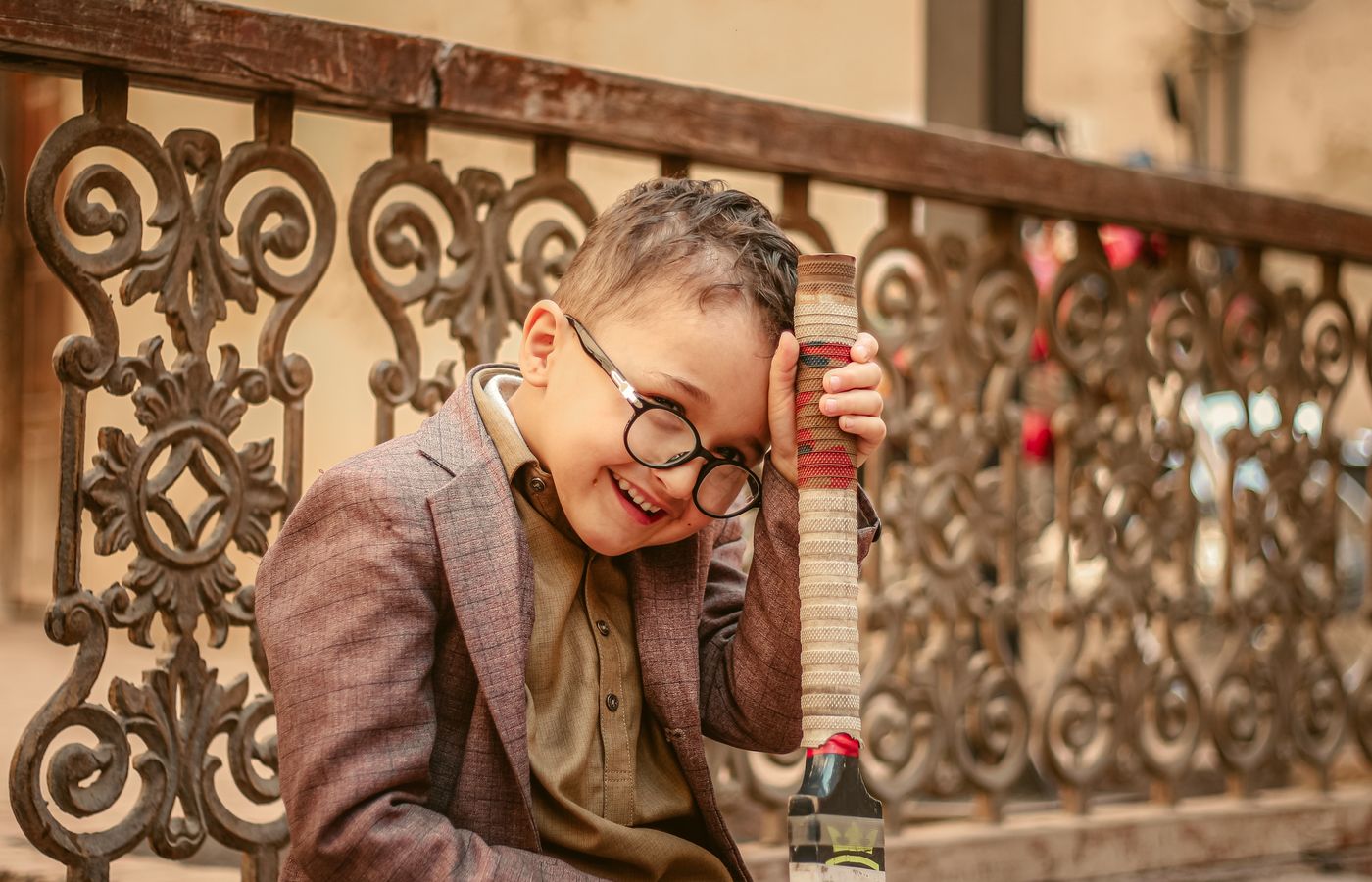 A joyful child with glasses wearing a brown suit, holding a toy in an outdoor setting.