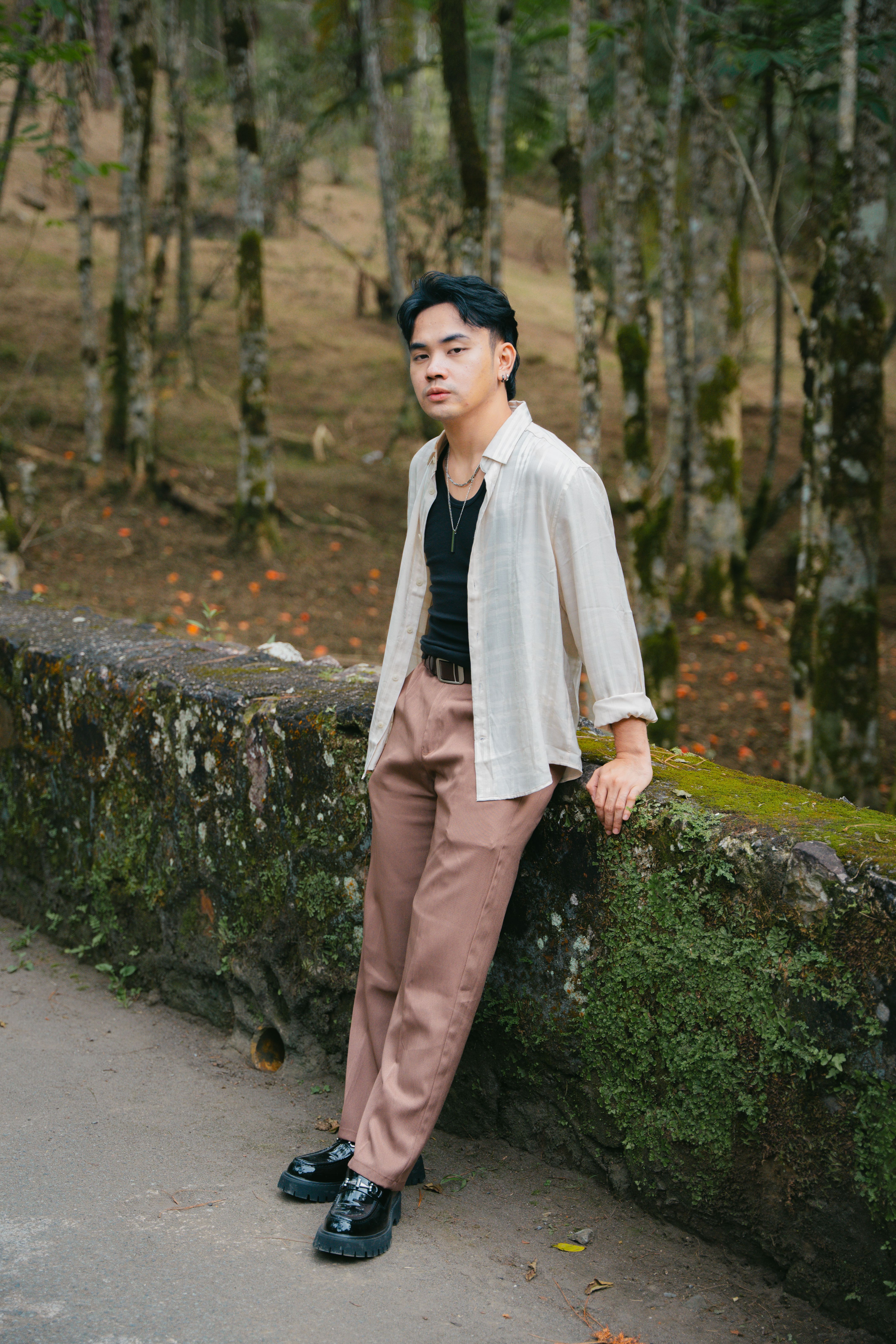 A fashionable young adult leans casually against a moss-covered wall in a serene forest scene.