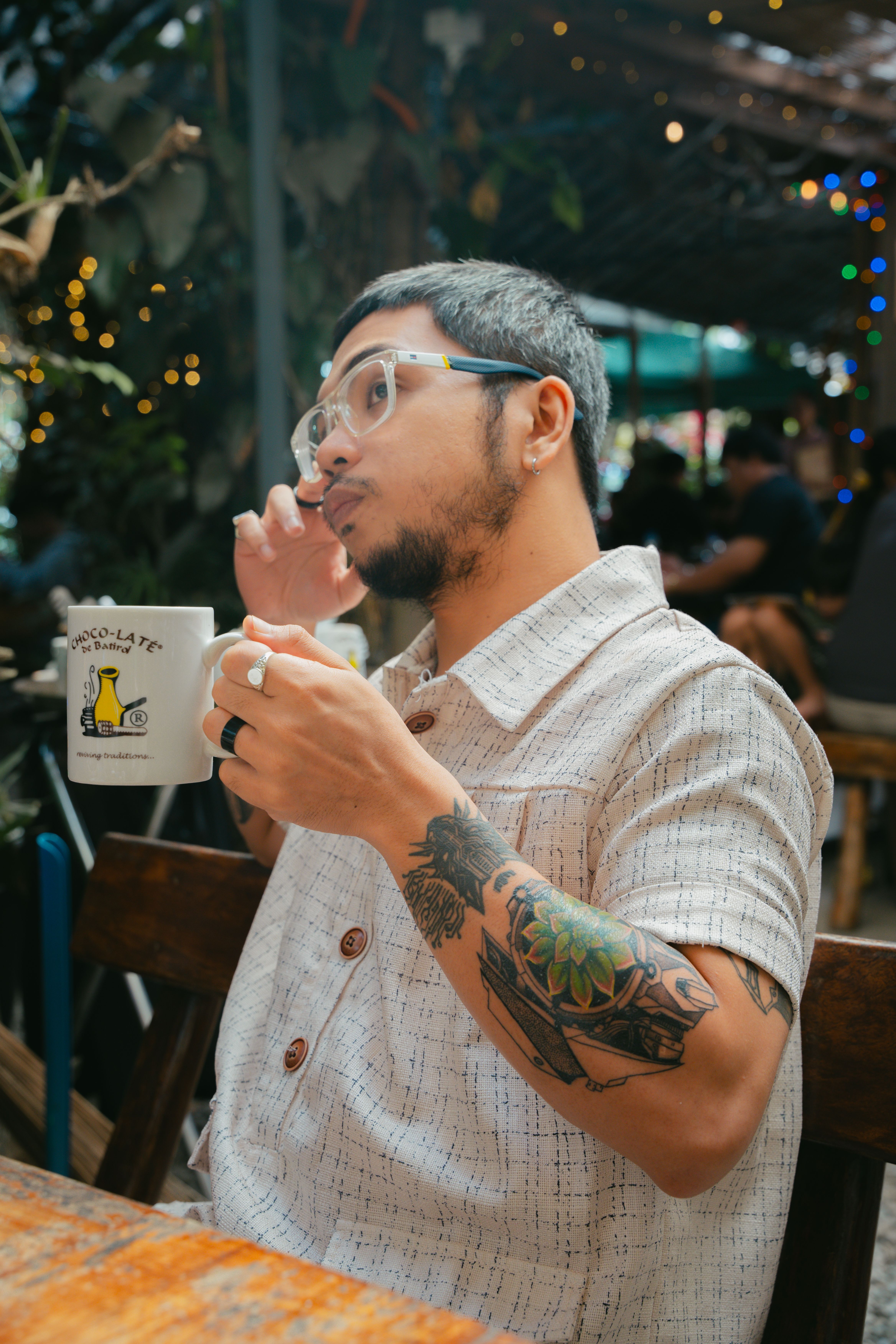 A tattooed man enjoys coffee in a lively outdoor cafe setting with festive lights.