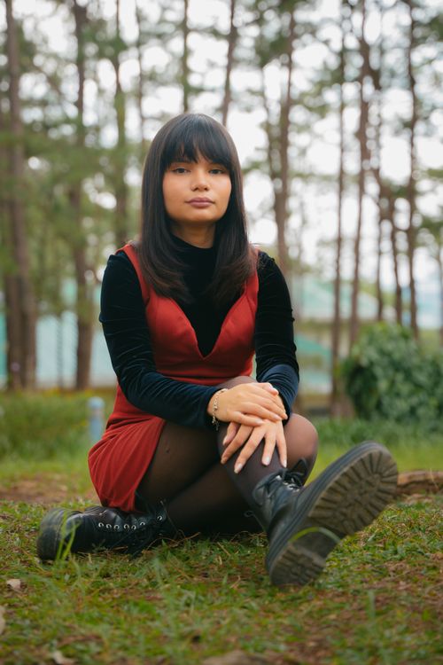 Free Young woman sitting in a forest, wearing a red dress and black boots, serene expression. Stock Photo