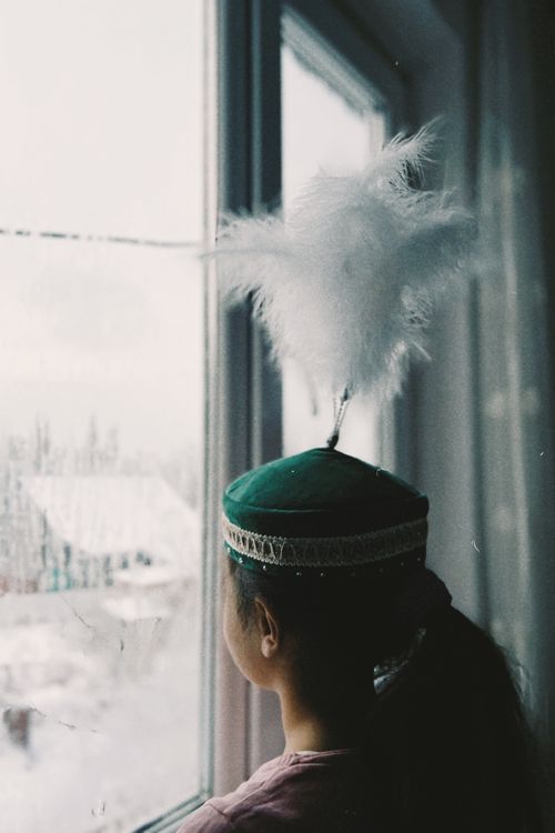 Free Young girl wearing a distinctive Kazakh hat, looking out snow-covered window in Talgar. Stock Photo