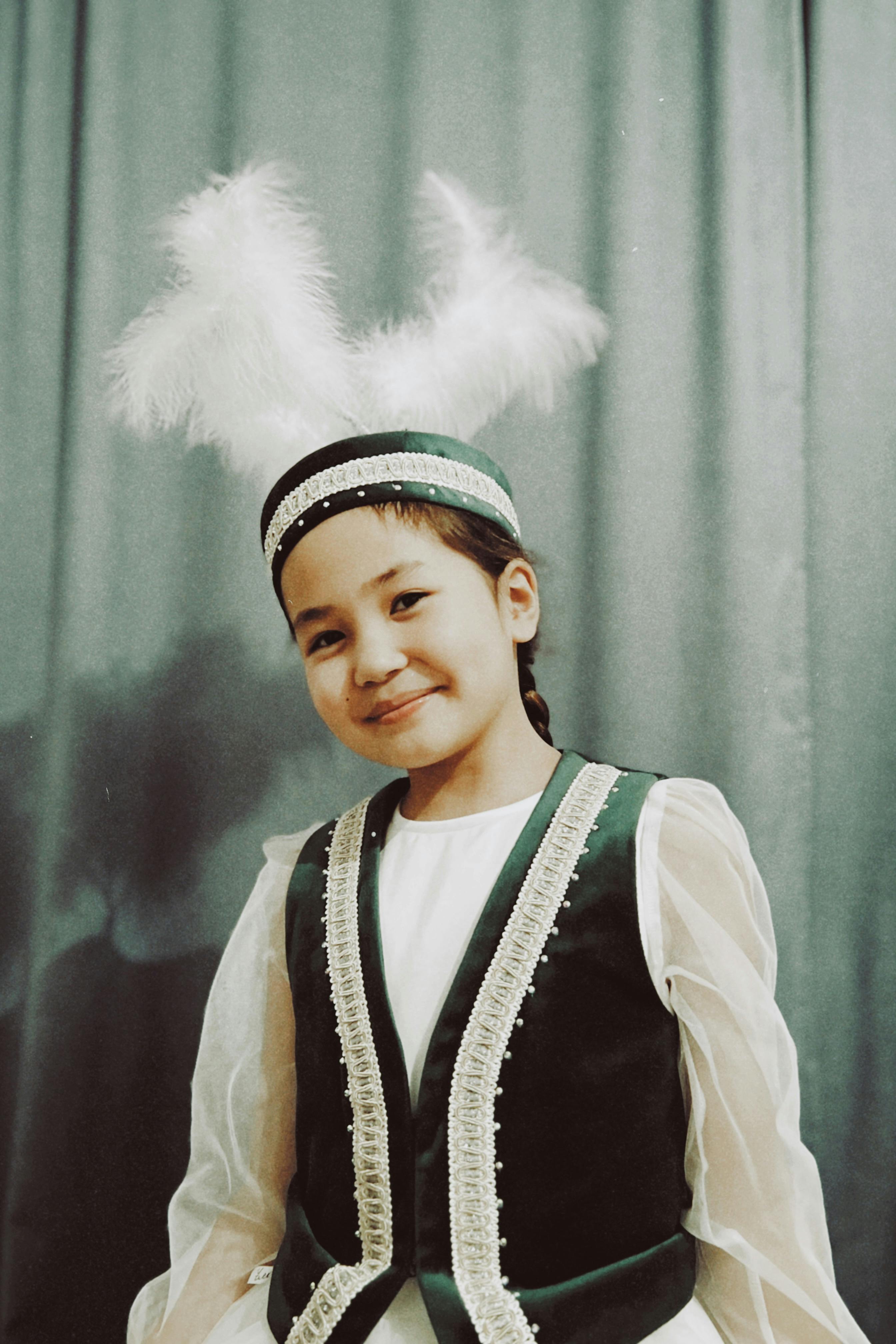 A young girl wearing traditional Kazakh clothing, with a feathered headdress, smiles warmly indoors.