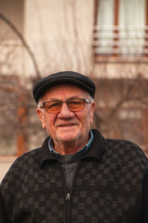 Free A senior man wearing glasses and a flat cap smiles warmly outdoors. Stock Photo
