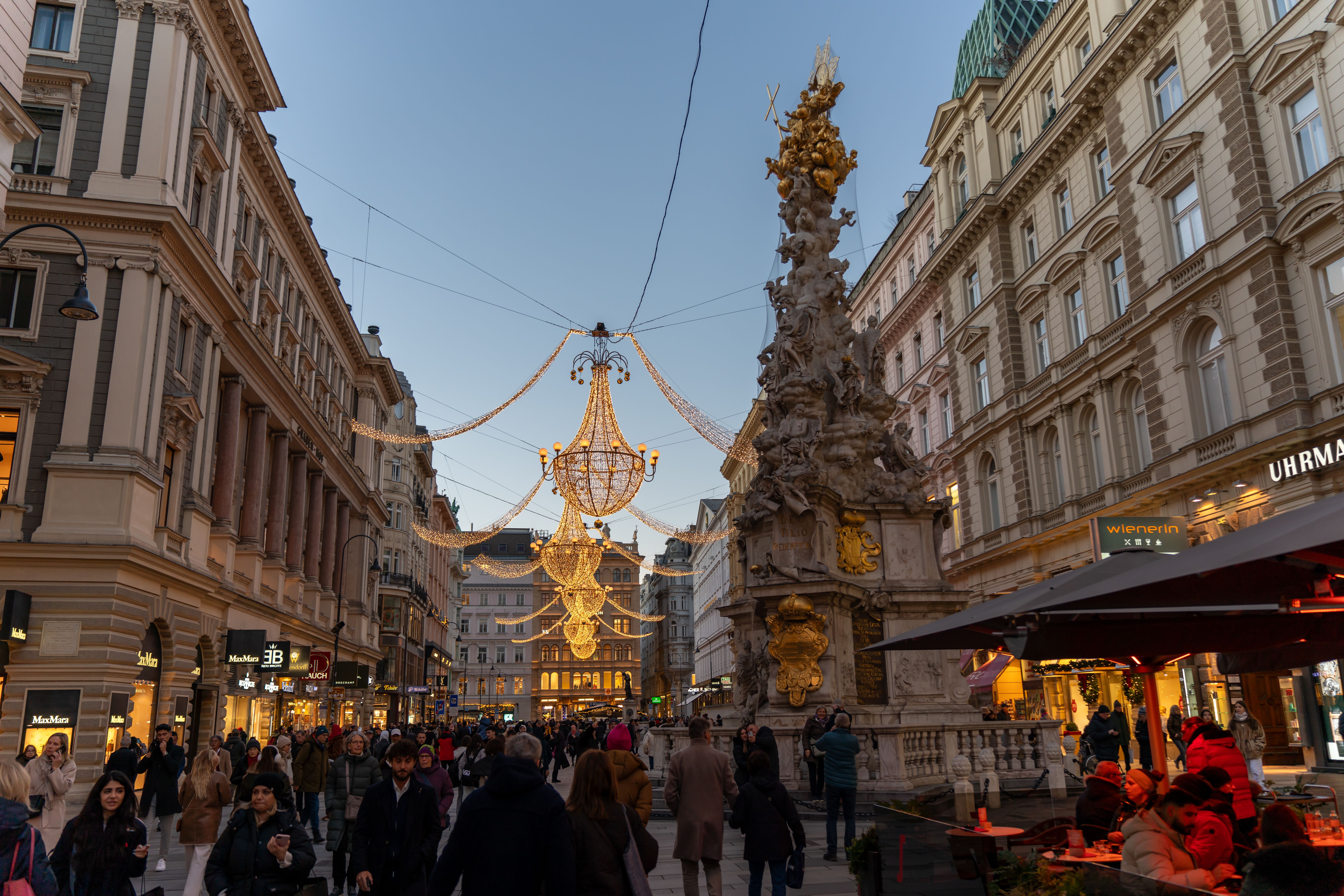 Lively Vienna street scene at twilight with holiday lights — Graben pedestrian spine + 1693 Pestsäule + Kohlmarkt to Hofburg