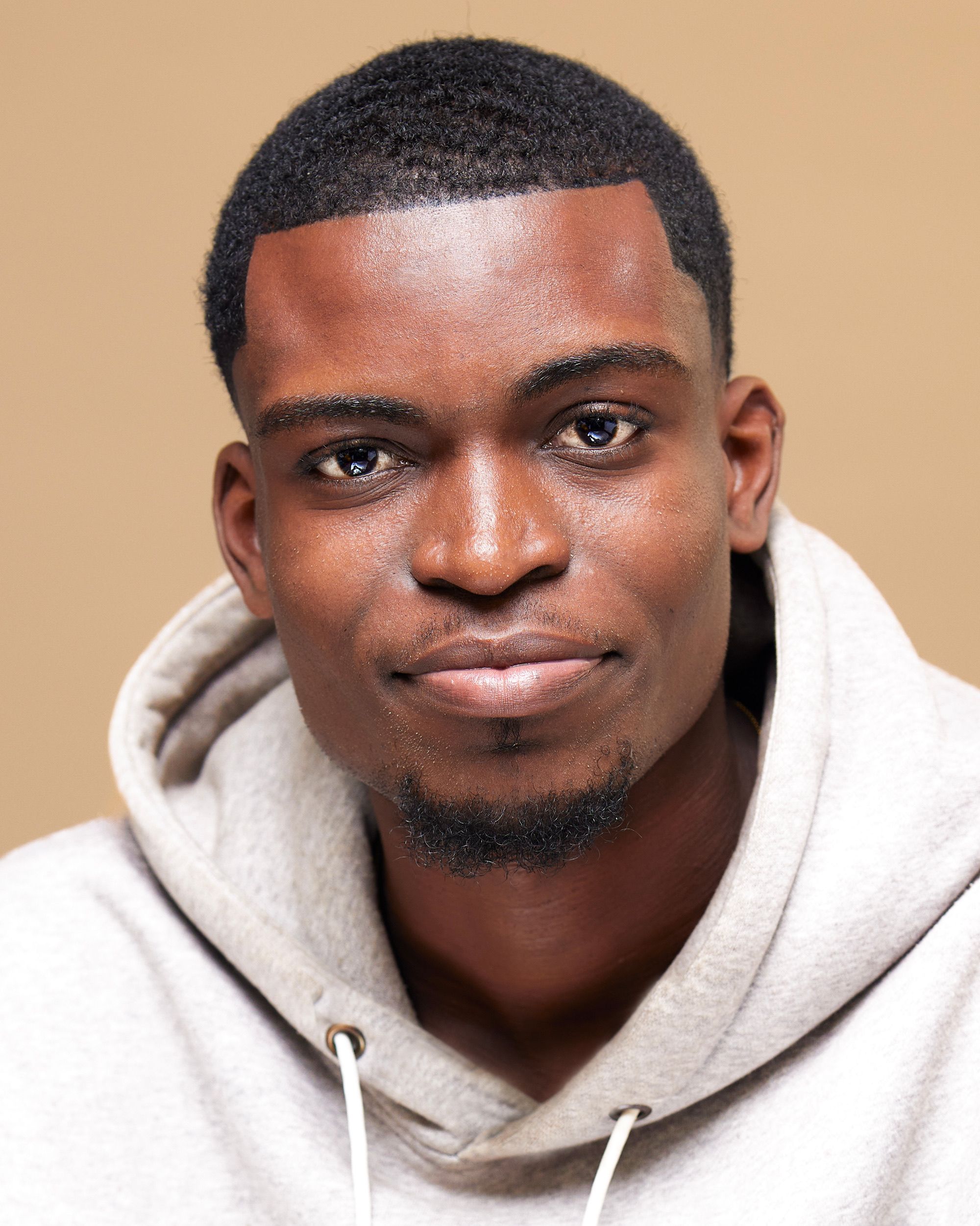 Close-up portrait of a young man smiling against a neutral background, wearing a casual hoodie.