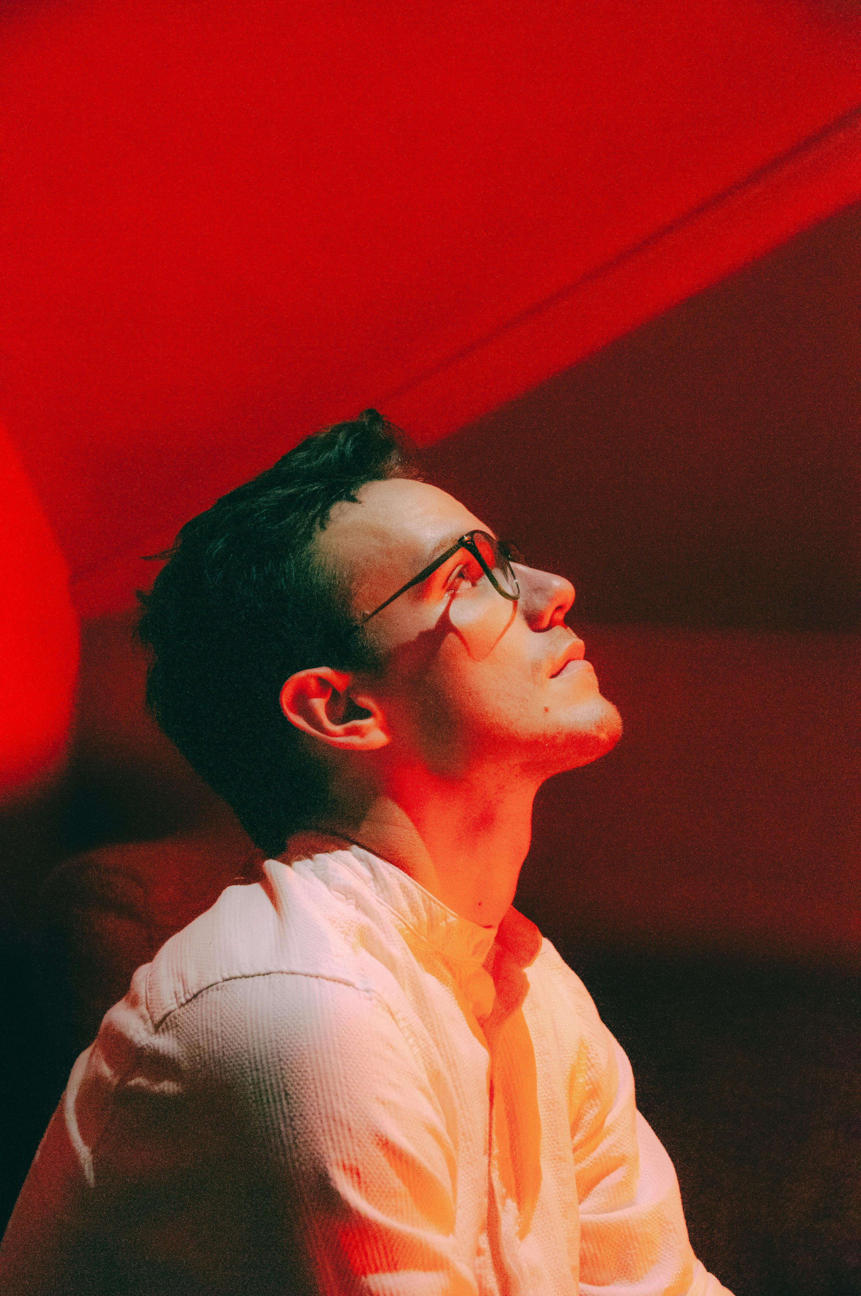 Portrait of a young man in glasses looking contemplative under red lighting.