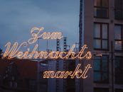 Glistening Christmas market sign lights up the evening sky in a city center, creating a warm festive ambiance.