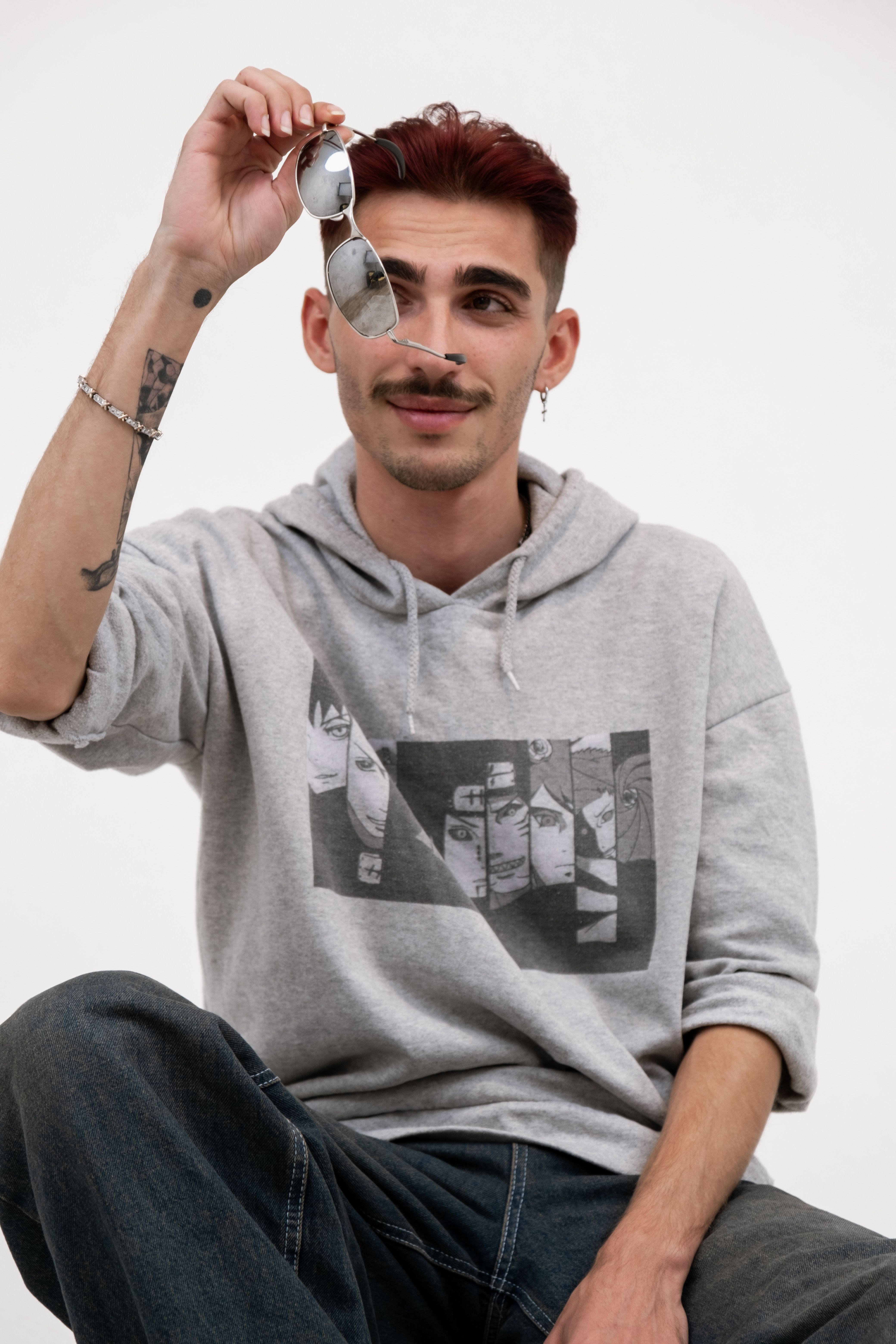 Young man holding sunglasses, wearing a graphic hoodie and jeans, styled with tattoos.
