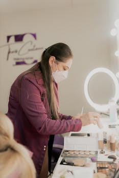 Makeup artist wearing mask arranging cosmetics in studio with illuminated mirror.