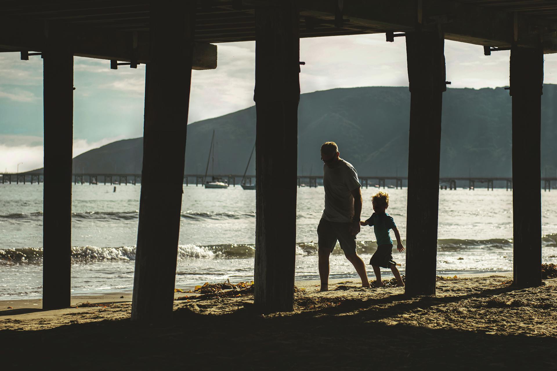 A father and son hold hands while walking on the beach under the pier at Avila Beach, CA.