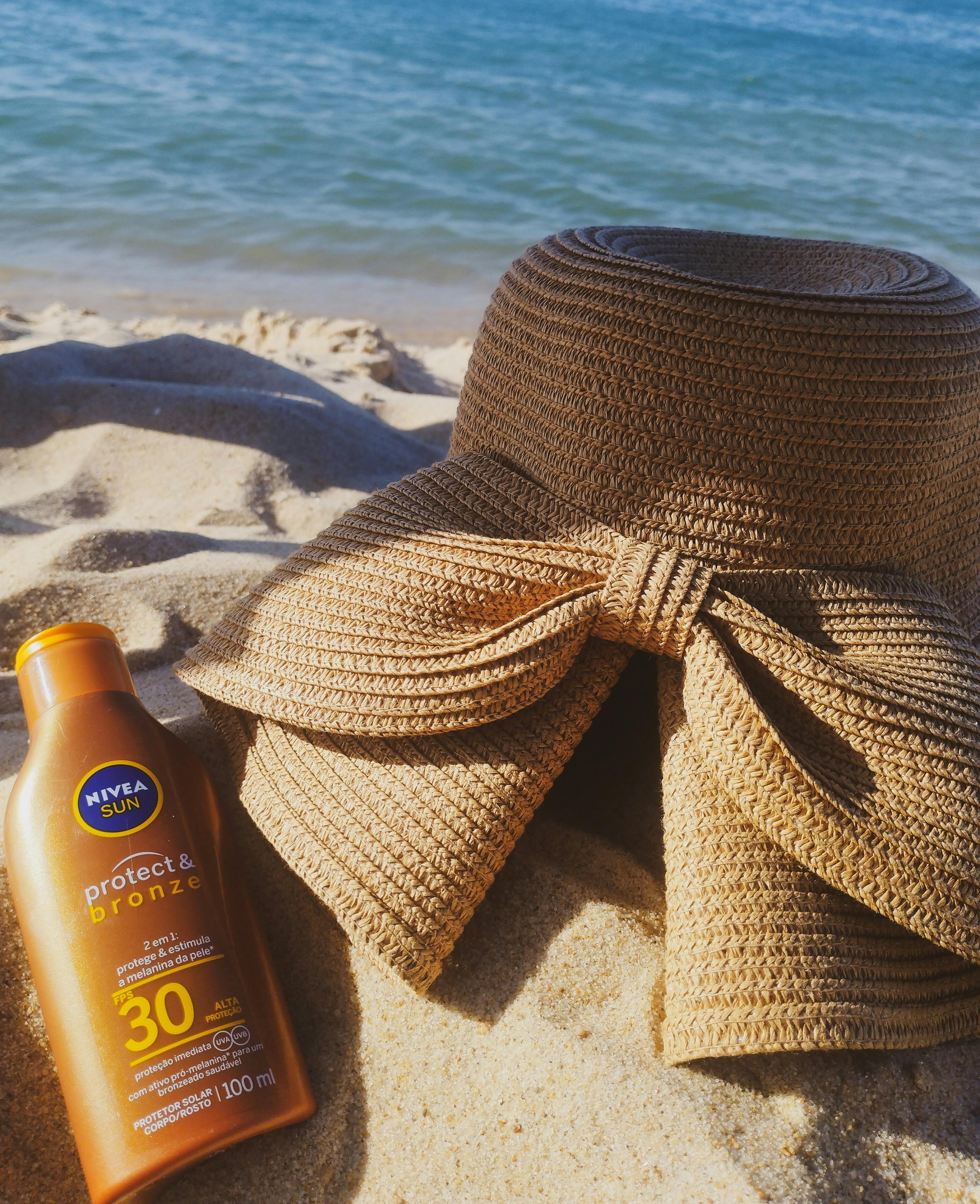 A sun hat and sunscreen on a sandy beach by the ocean, perfect for summer.