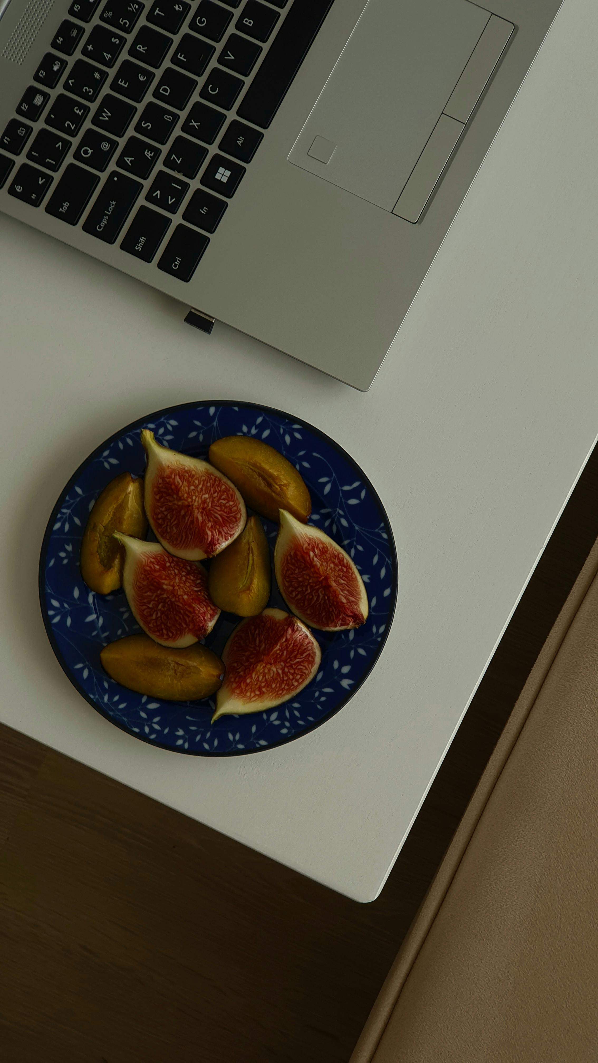 Aerial view of figs and plums on a plate near a laptop on a white desk, perfect for healthy snacking inspiration.