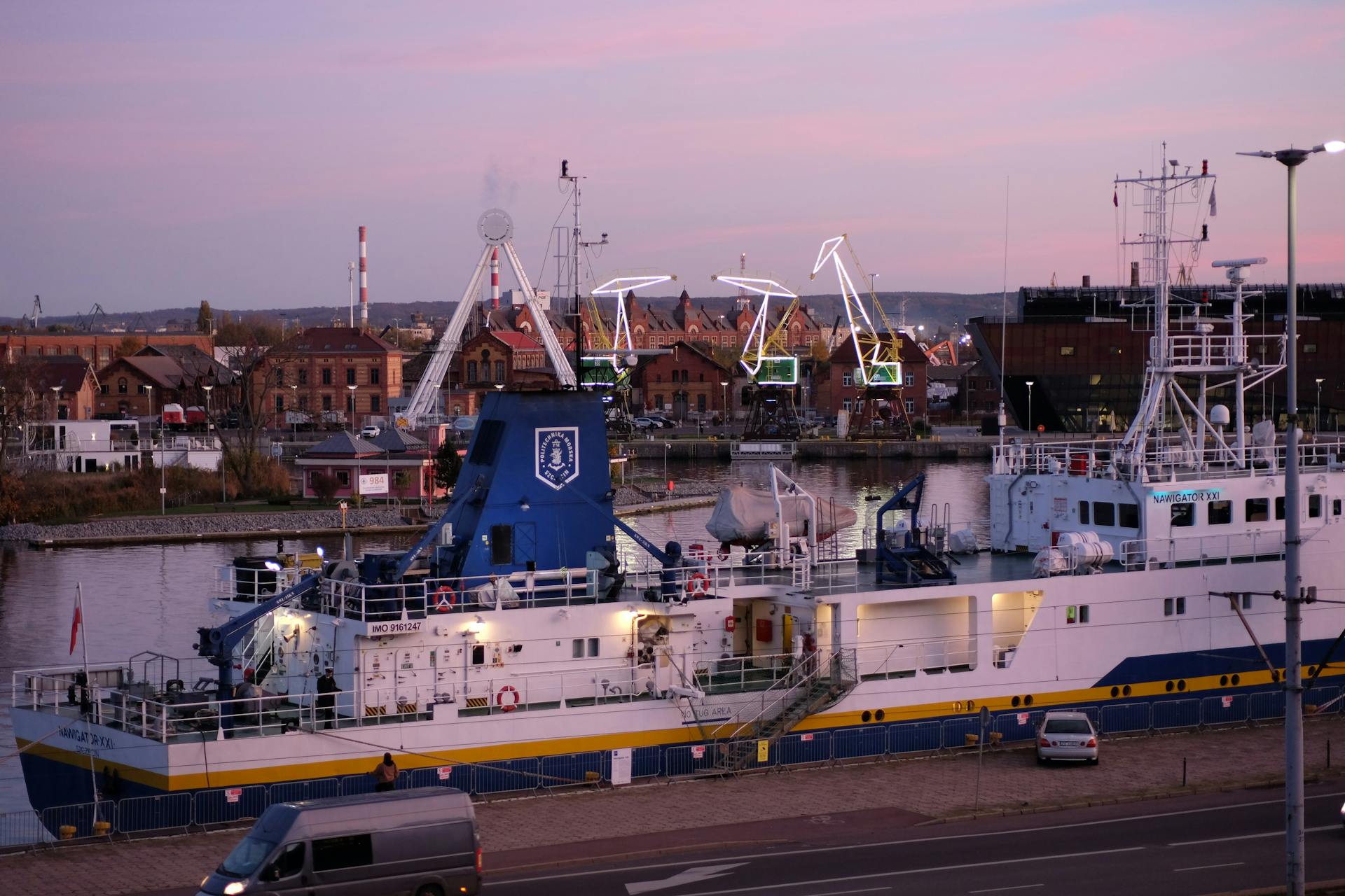 A large industrial ship docked at the harbor with cranes and buildings in the background during twilight.