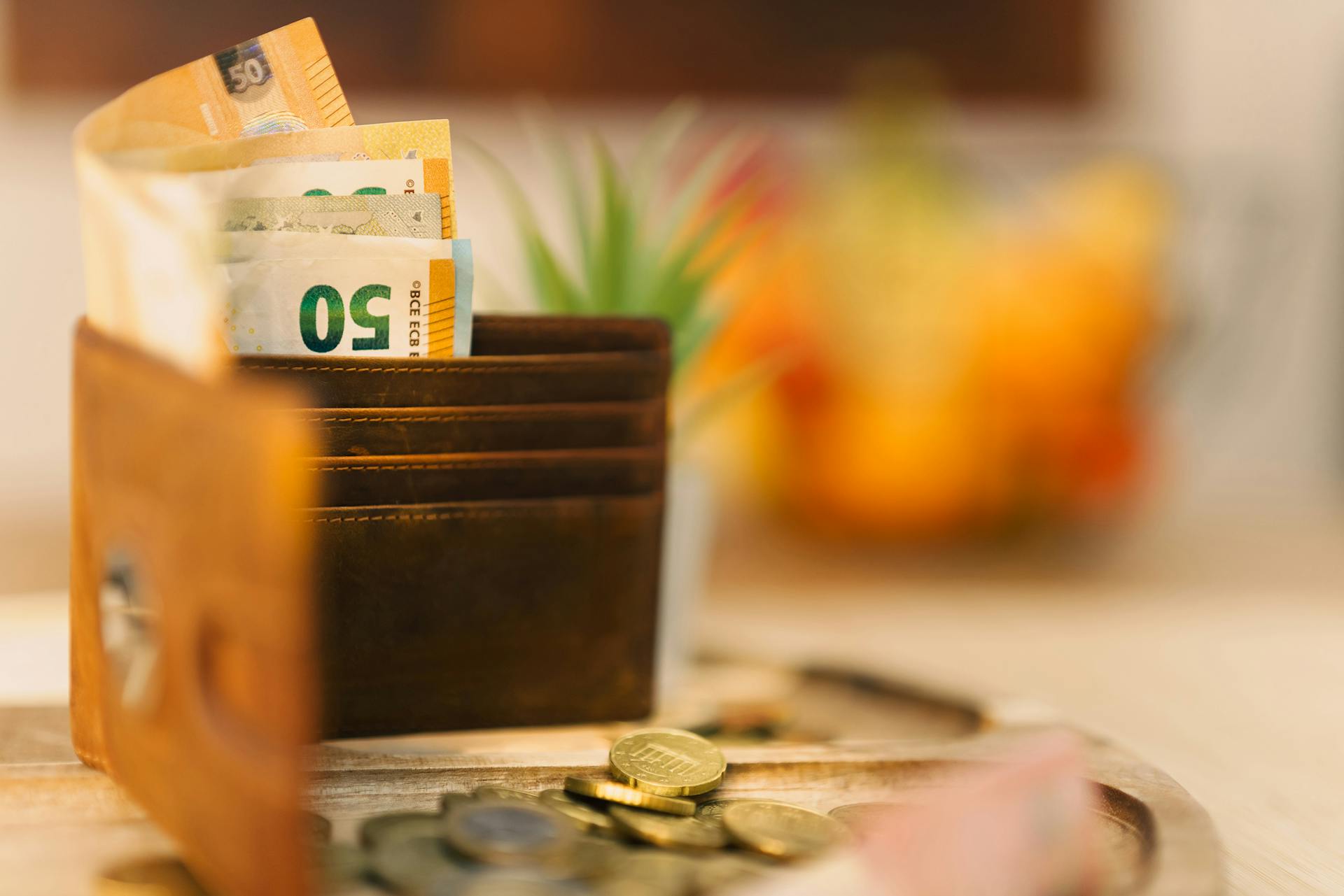 Brown wallet holding banknotes with scattered coins on a wooden table.