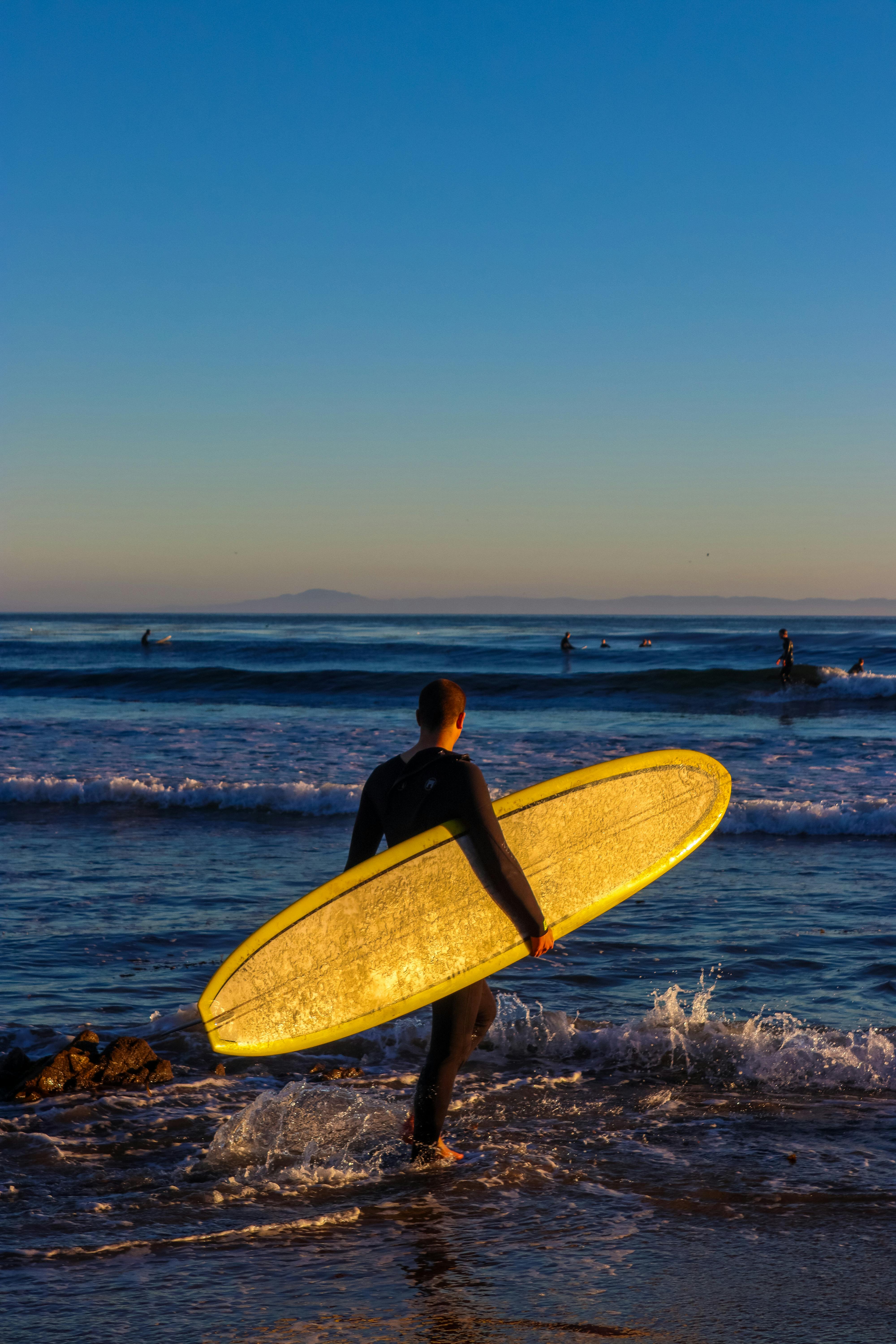 A surfer with a yellow surfboard heads into the ocean at sunset in Santa Barbara.