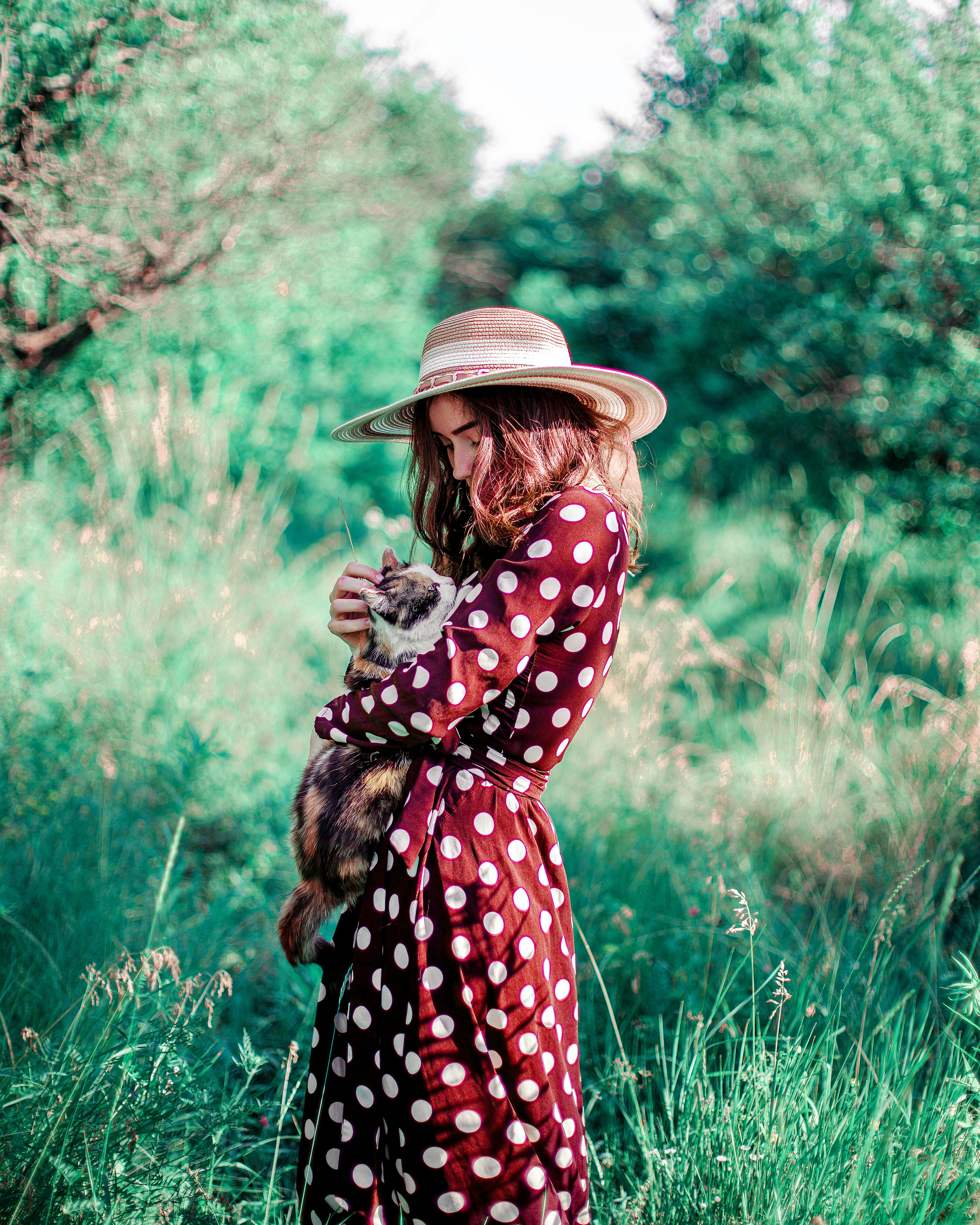 Young woman holding a fluffy cat in a vibrant green field, wearing a polka dot dress and sun hat.