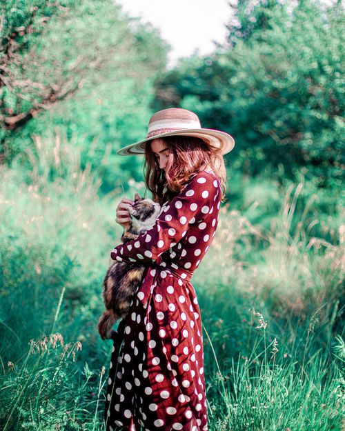 Free Young woman holding a fluffy cat in a vibrant green field, wearing a polka dot dress and sun hat. Stock Photo