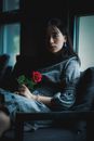 在室内柔和的灯光下，一位年轻女子手拿一朵红玫瑰，沉思地凝视着