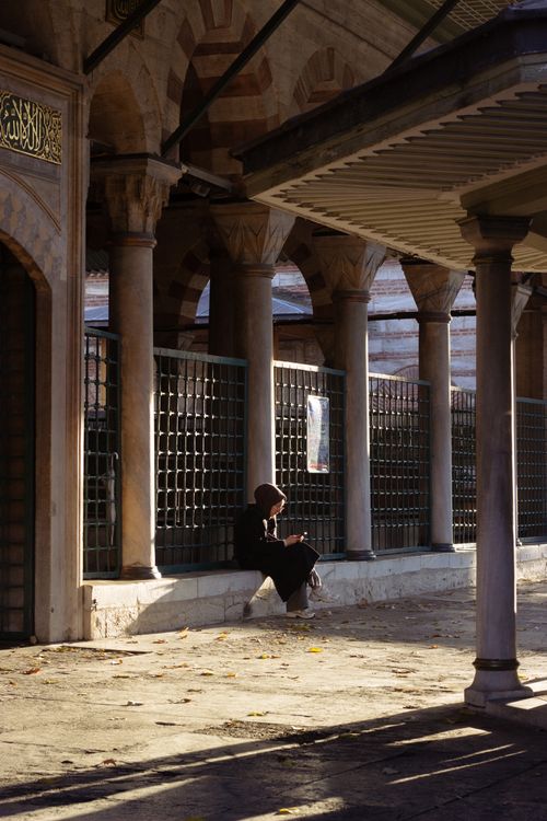 Free Woman sitting in a historic mosque courtyard with pillars casting long shadows in sunlight. Stock Photo