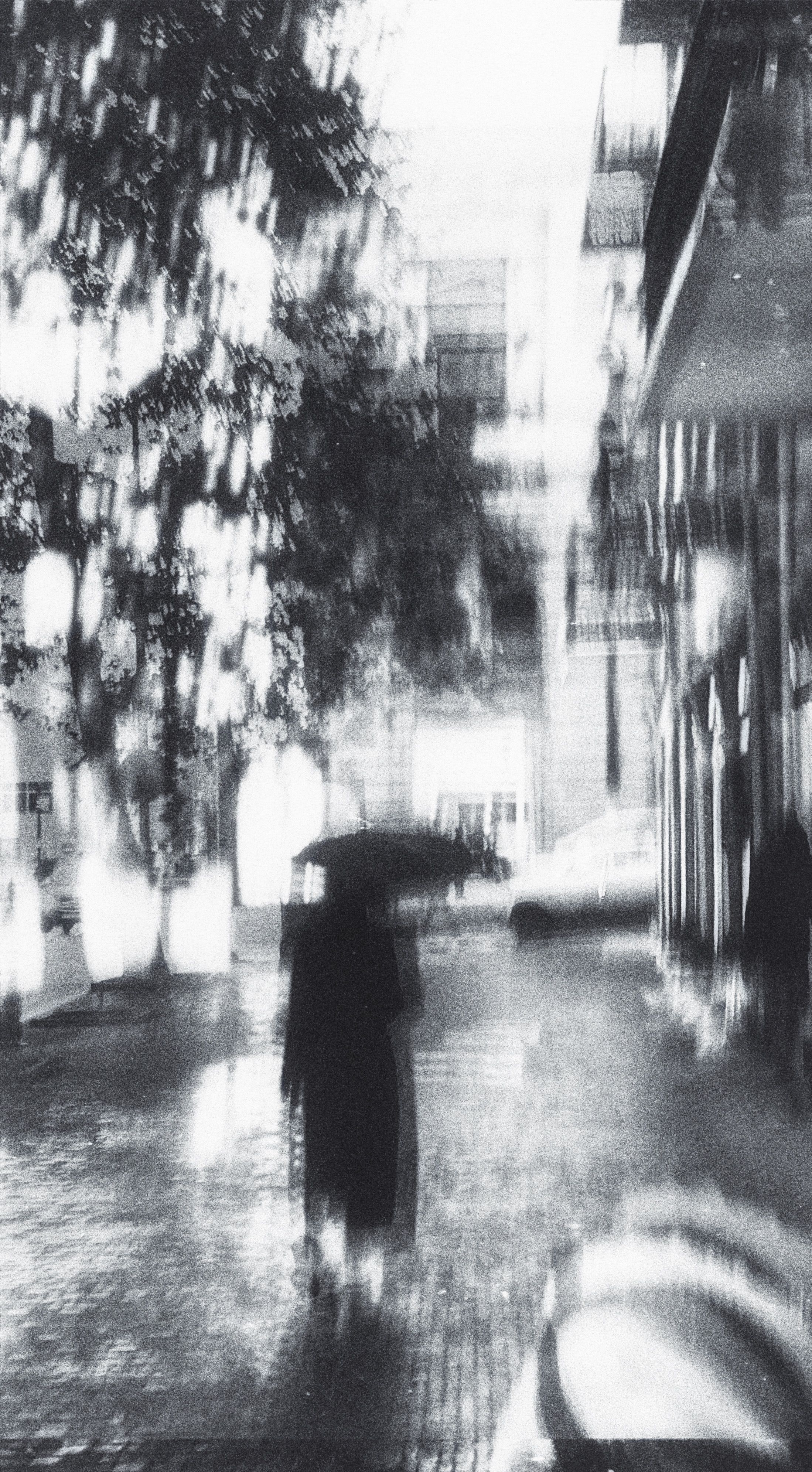 A moody, monochrome scene of a street during rainfall with a blurred figure holding an umbrella.