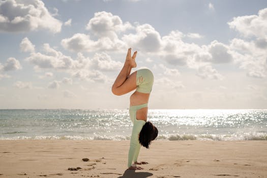 A woman in a handstand yoga pose on a serene beach under a bright sky.