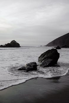 A serene coastal scene featuring dark sea rocks and gentle waves under a cloudy sky.