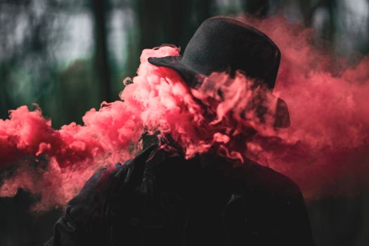 A mysterious figure in a hat surrounded by vibrant red smoke outdoors.