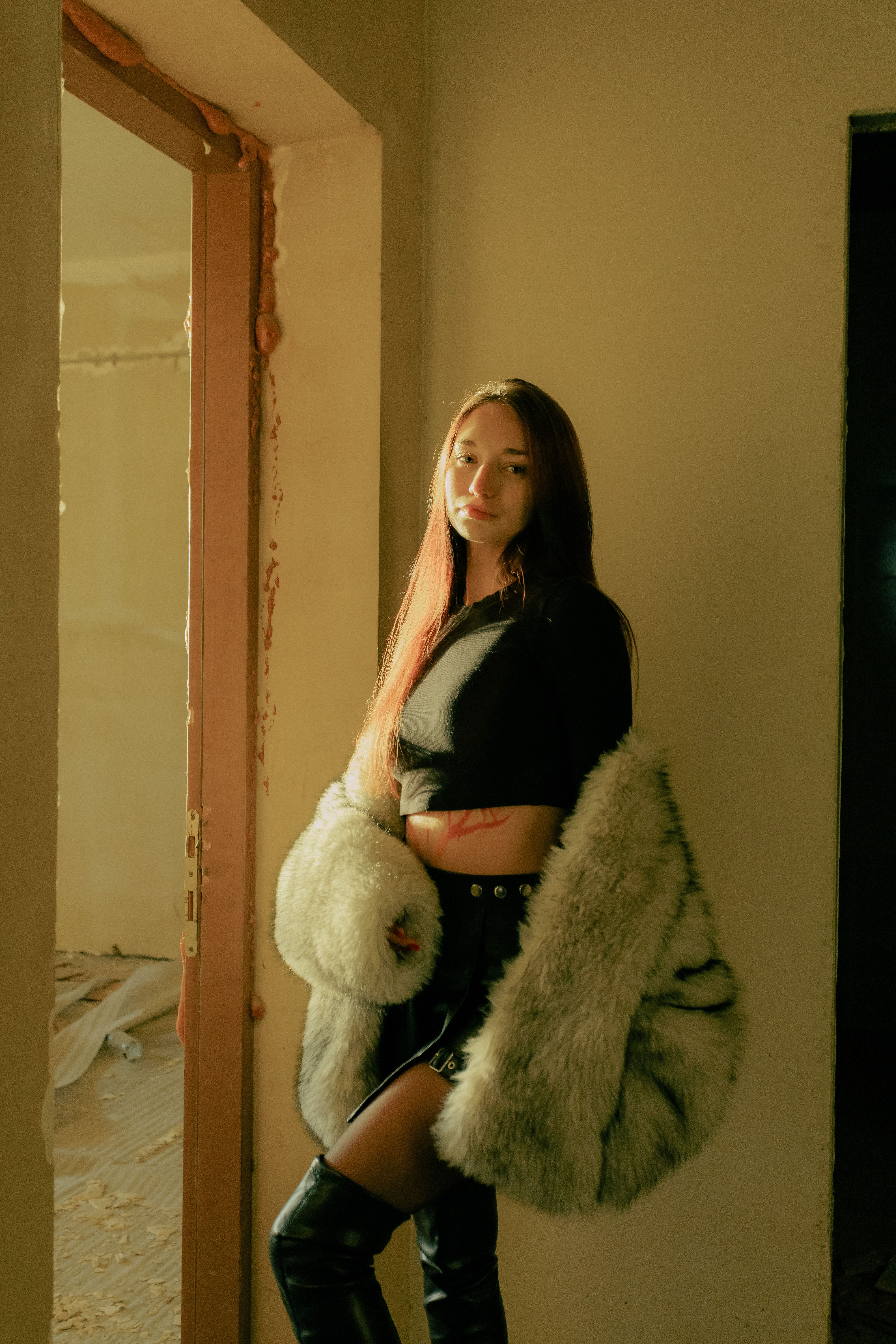 Elegant woman in black and fur coat posing in sunlit indoor setting.