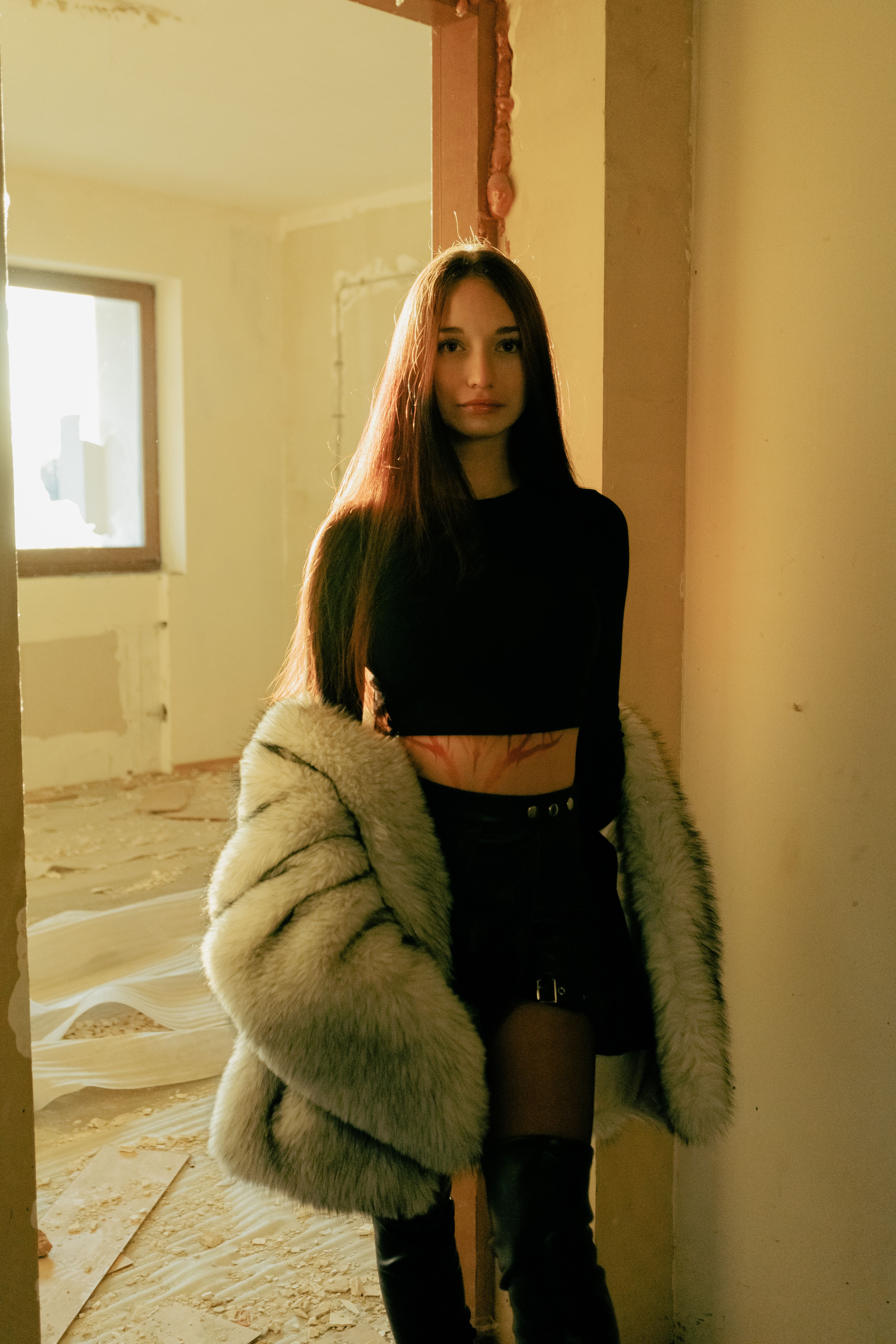 Elegant woman in fur coat standing in a sunlit abandoned interior.