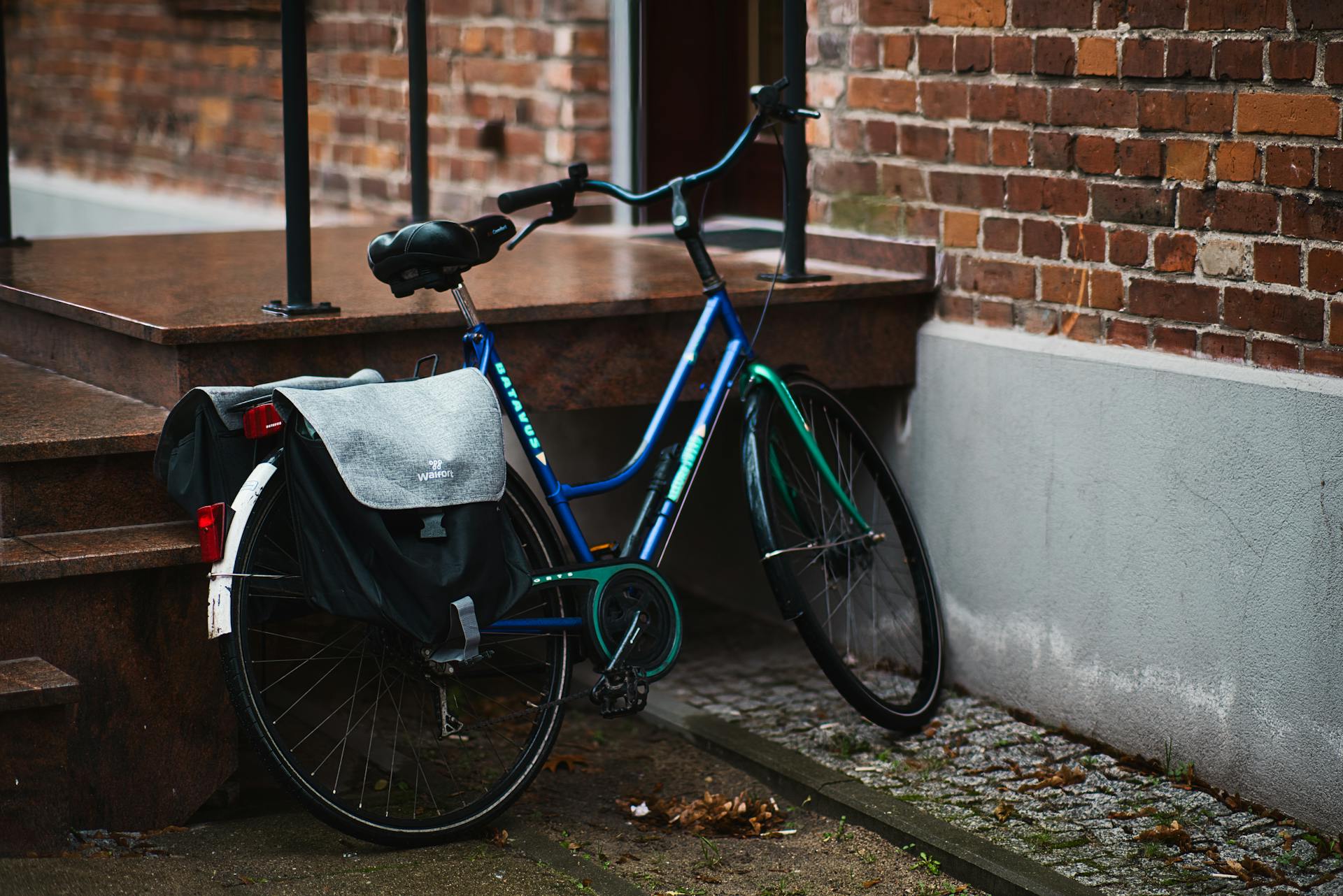 A blue bicycle with panniers rests against a brick wall in an urban setting.