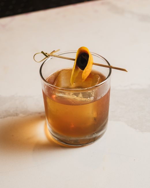 A bourbon Old Fashioned in a rocks glass over one large ice cube, garnished with an orange twist and a cocktail cherry, with a bottle of bourbon in the background.