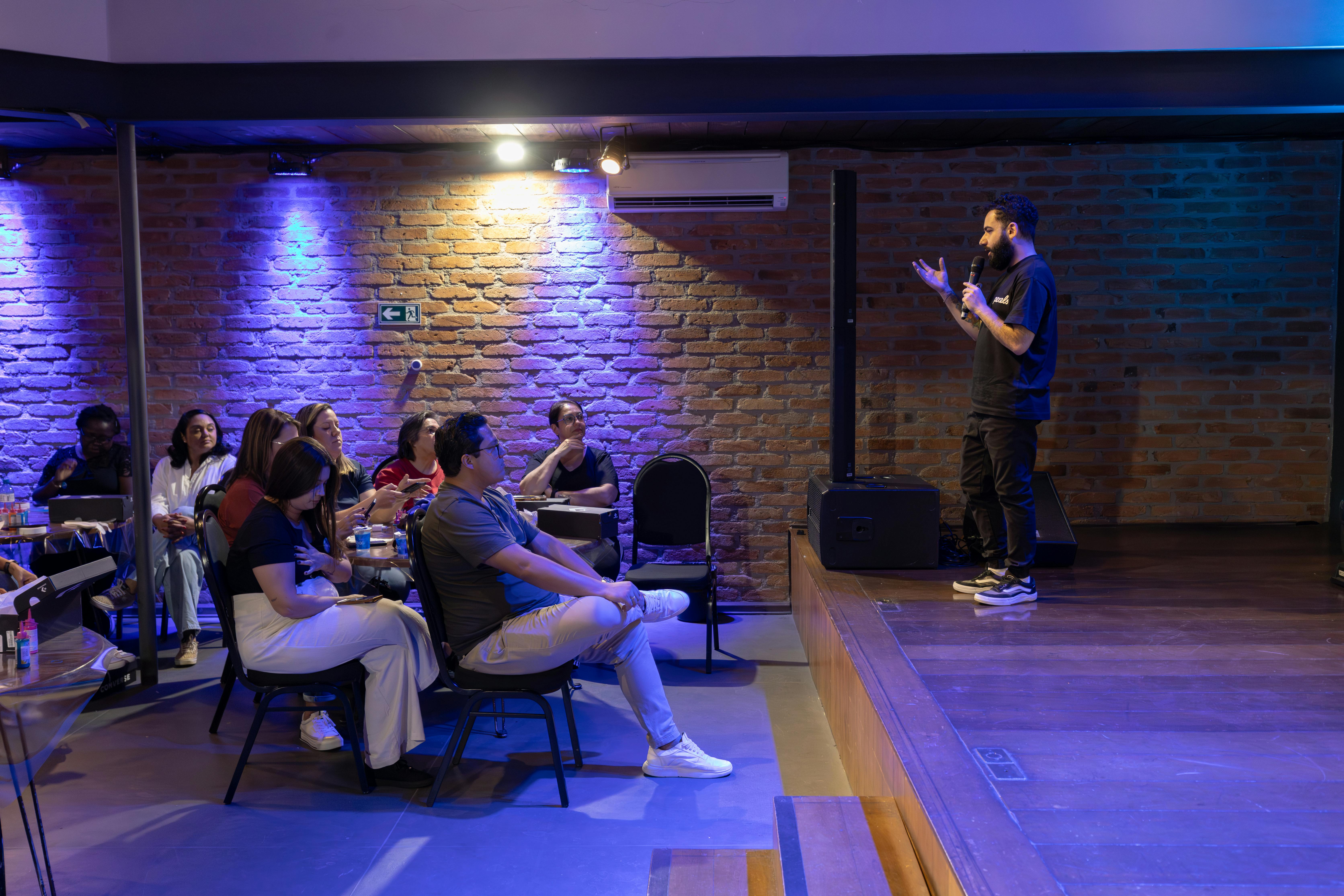 A speaker presents to an attentive audience in a modern indoor setting with brick walls and ambient lighting.