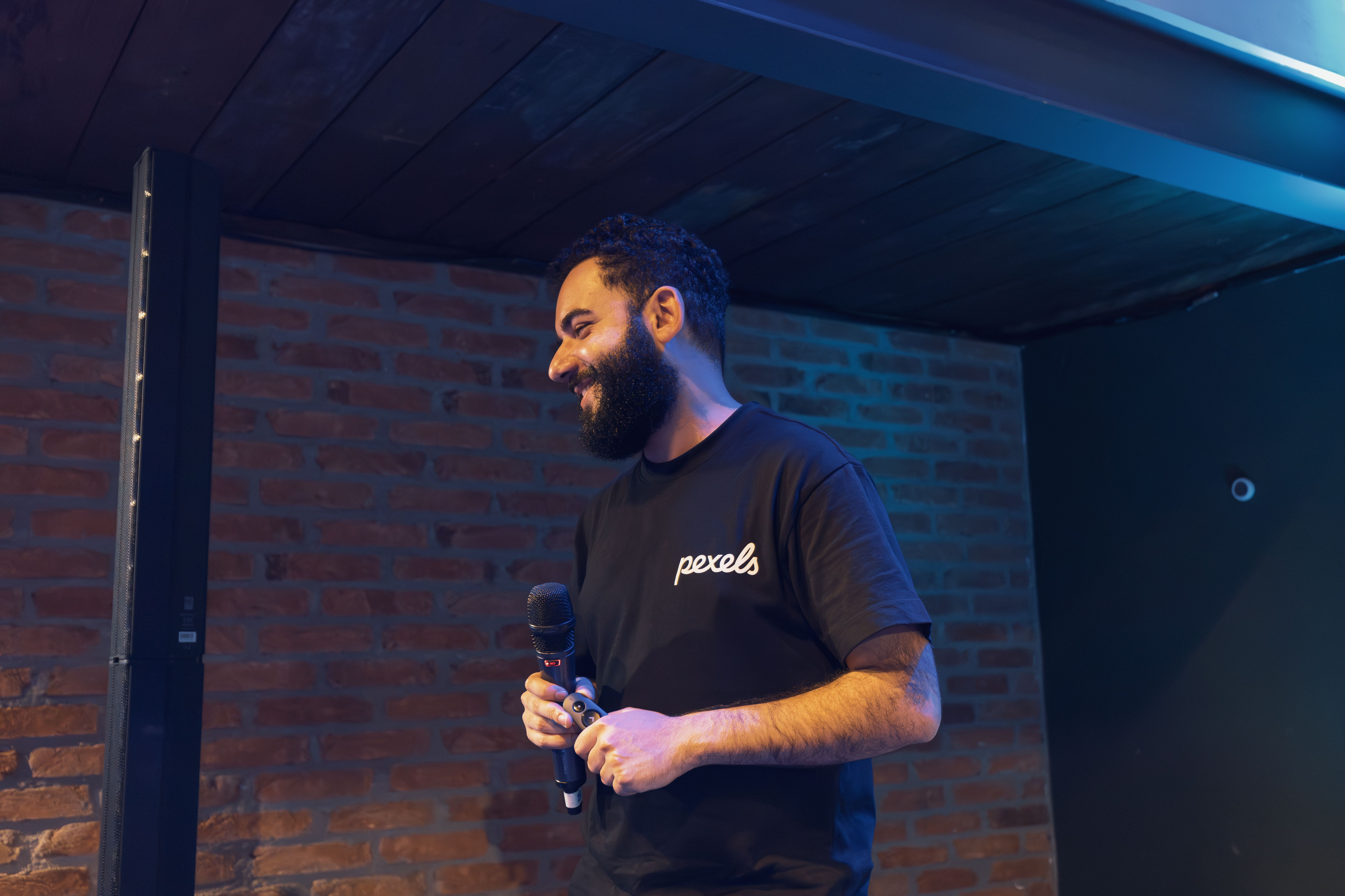 Engaging speaker at an indoor event with a microphone, wearing a Pexels shirt.