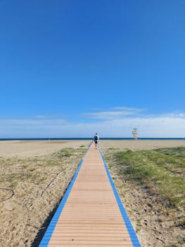 A lone figure walks on a wooden boardwalk toward the ocean under a clear blue sky.