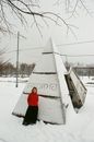 冬季的公园里，一名身穿红色毛衣的女子在白雪覆盖的金字塔结构旁摆着姿势