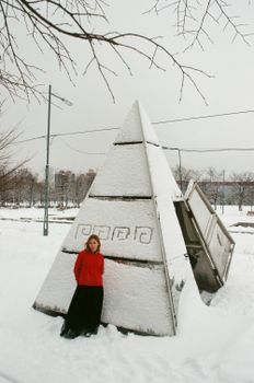 冬季的公园里，一名身穿红色毛衣的女子在白雪覆盖的金字塔结构旁摆着姿势