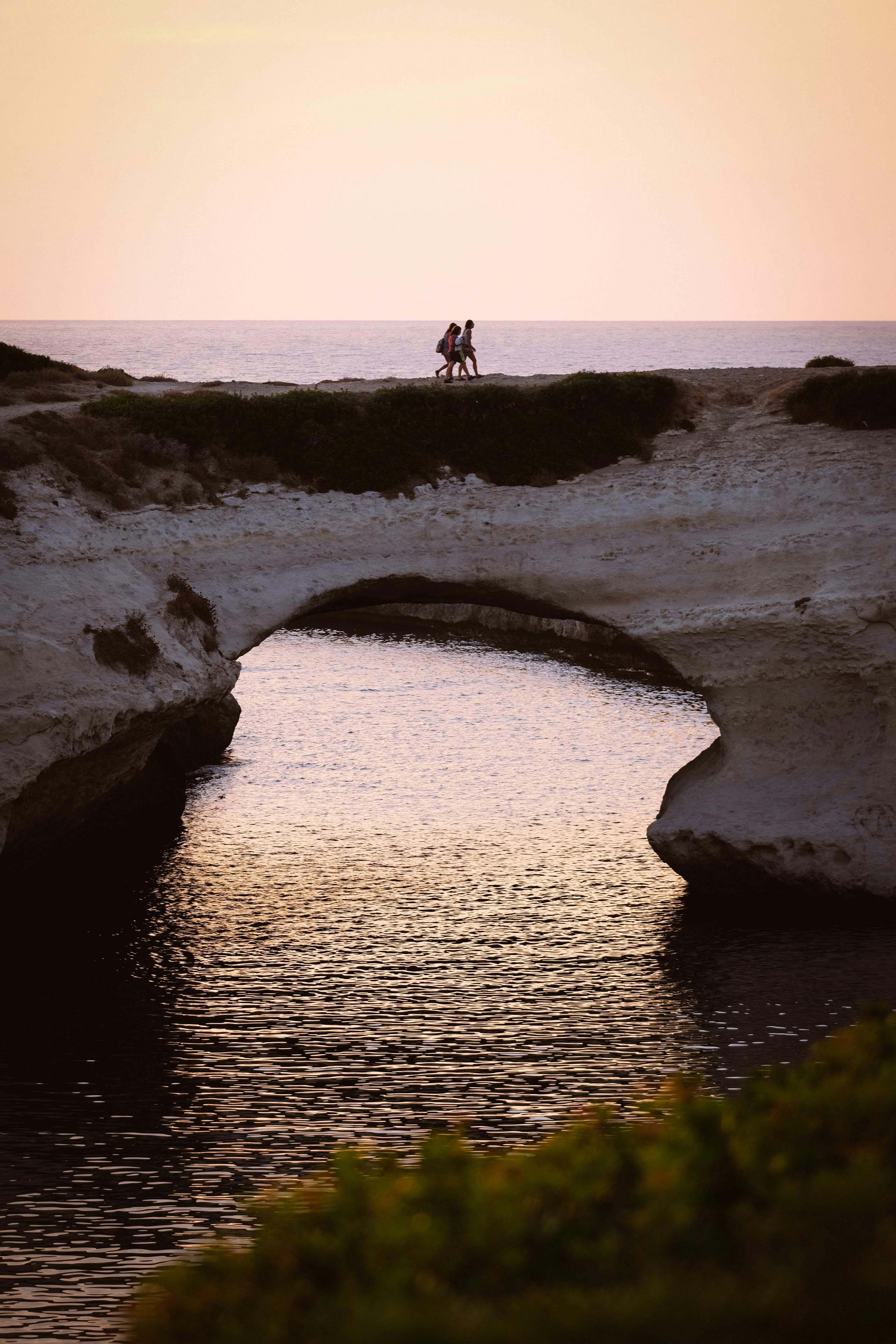 日落时分，一对夫妇在海边穿过天然石拱门，场景宁静