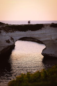 日落时分，一对夫妇在海边穿过天然石拱门，场景宁静
