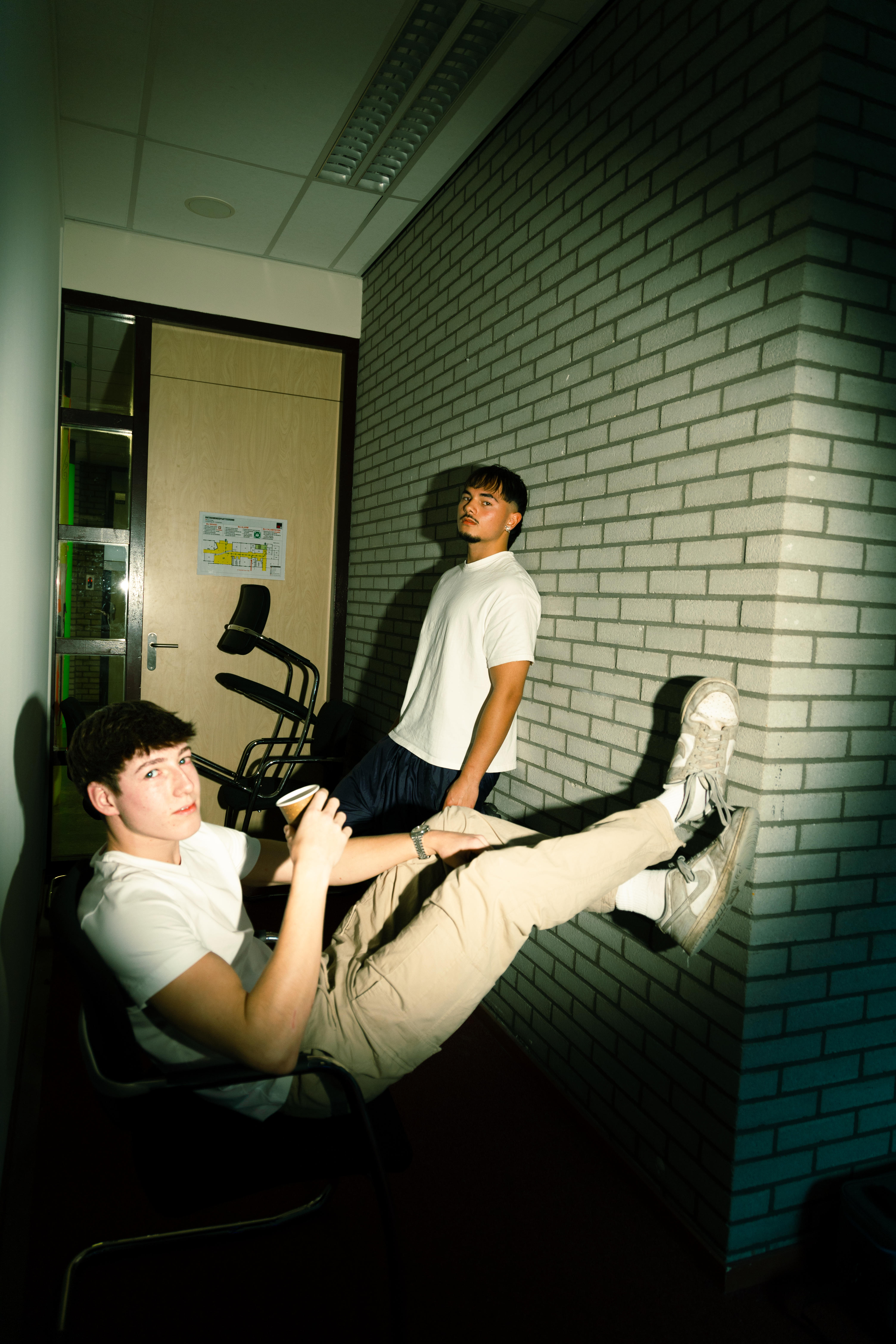 Two young men in relaxed poses indoors against a brick wall.