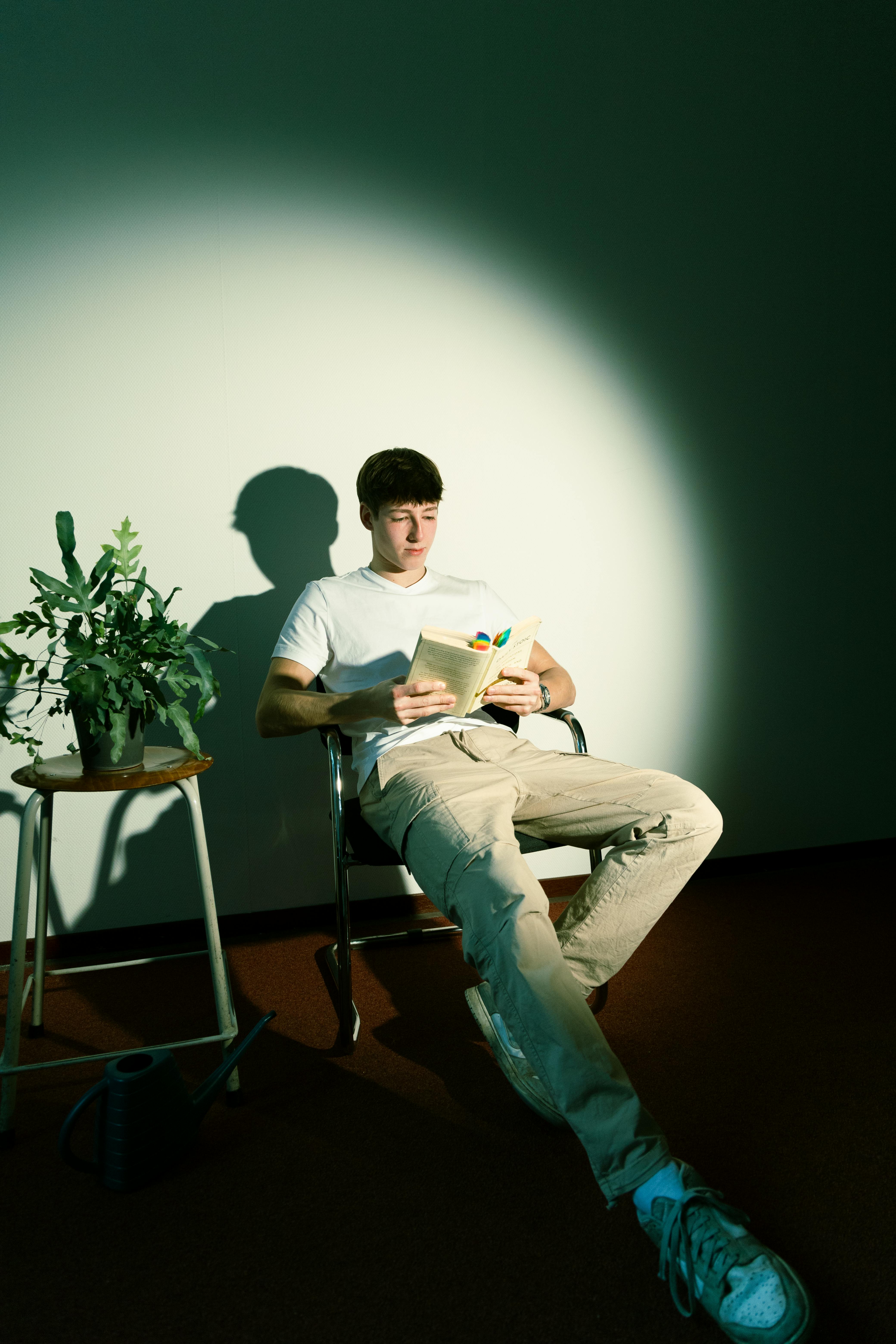 A young man sits reading in a spotlight beside a houseplant. Calm and focused atmosphere.