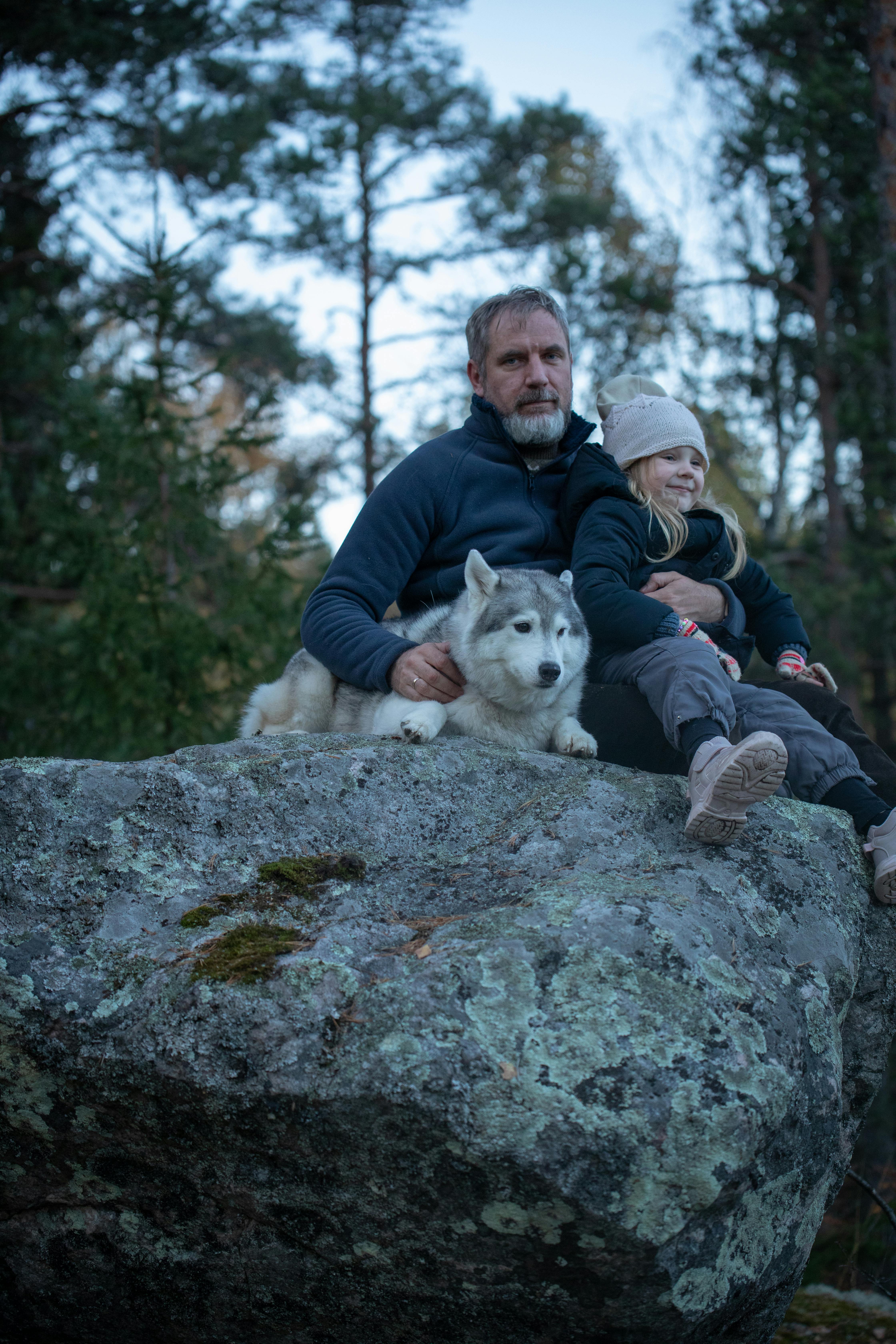 A father and daughter sit with a Husky on a rock in a serene forest setting.